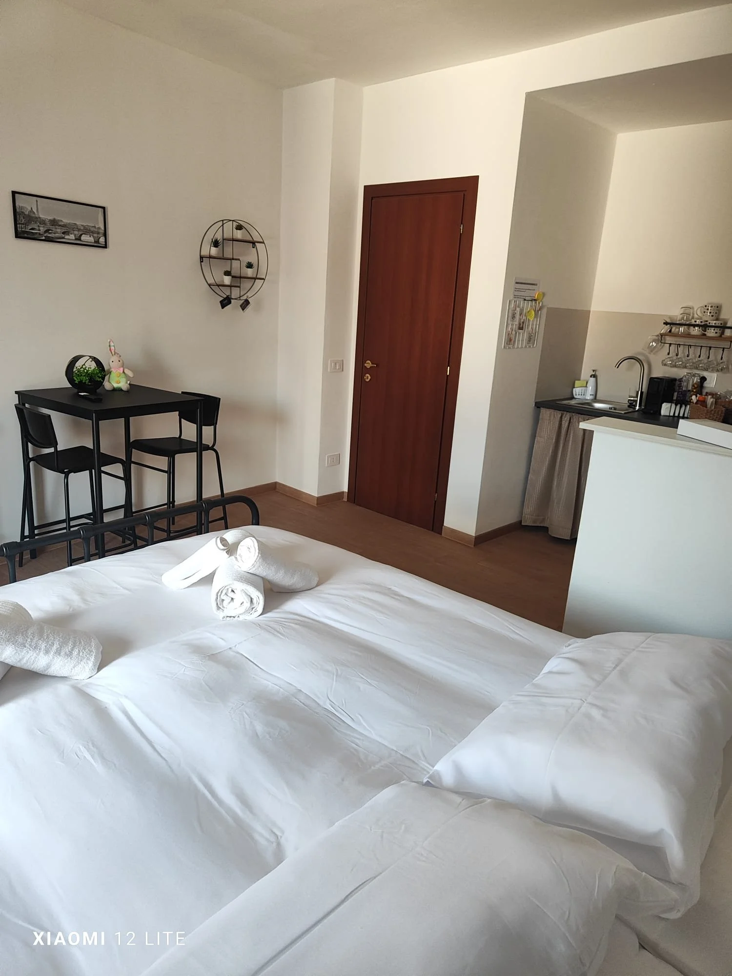 Camera da letto con letto coperto da lenzuola bianche, con asciugamani piegati a forma di animali sul letto, e arredamento minimalista come tavolo e sedie nere, pareti bianche, porta in legno, e angolo cucina con lavabo, dispenser di sapone, e scaffali con tazze e altri utensili.