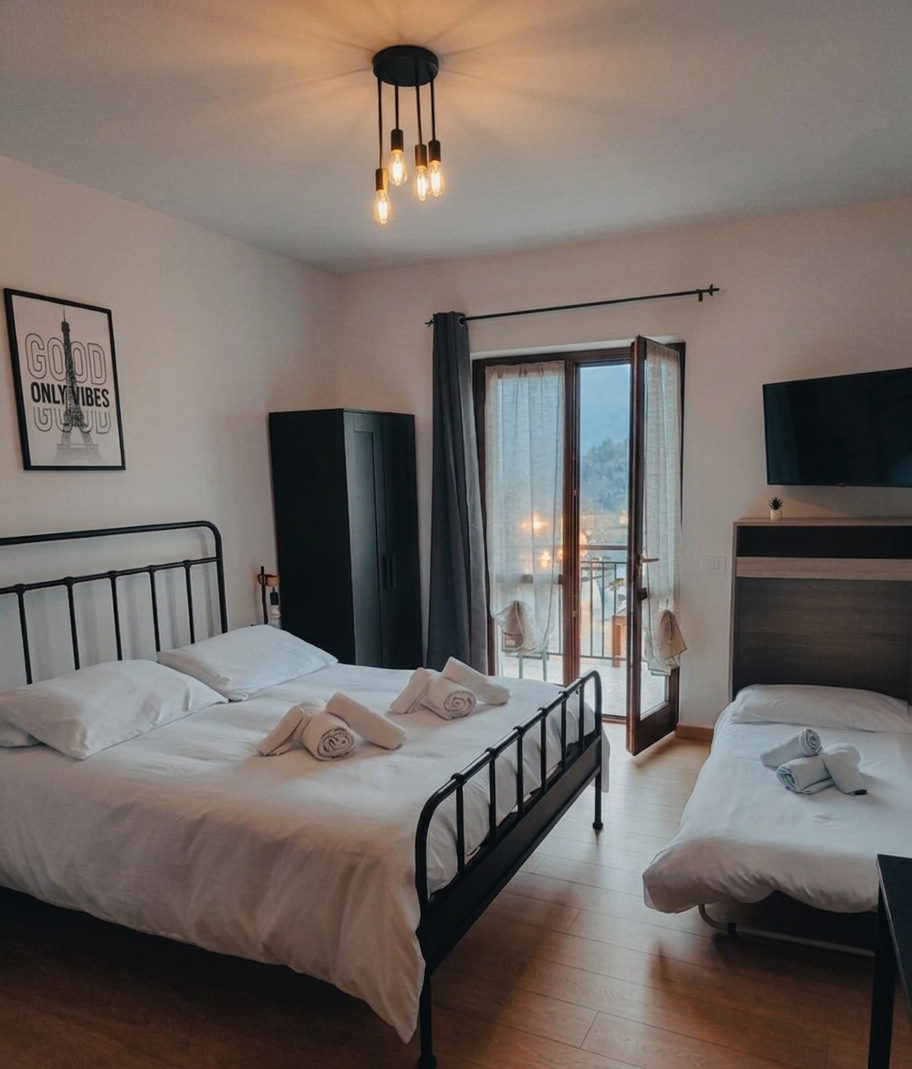 Camera da letto con un grande letto, un letto singolo, un armadio nero, una parete con quadro con scritta 'Good Vibes Only', balcone con vista sulla città, lampadario con lampadine esposte.