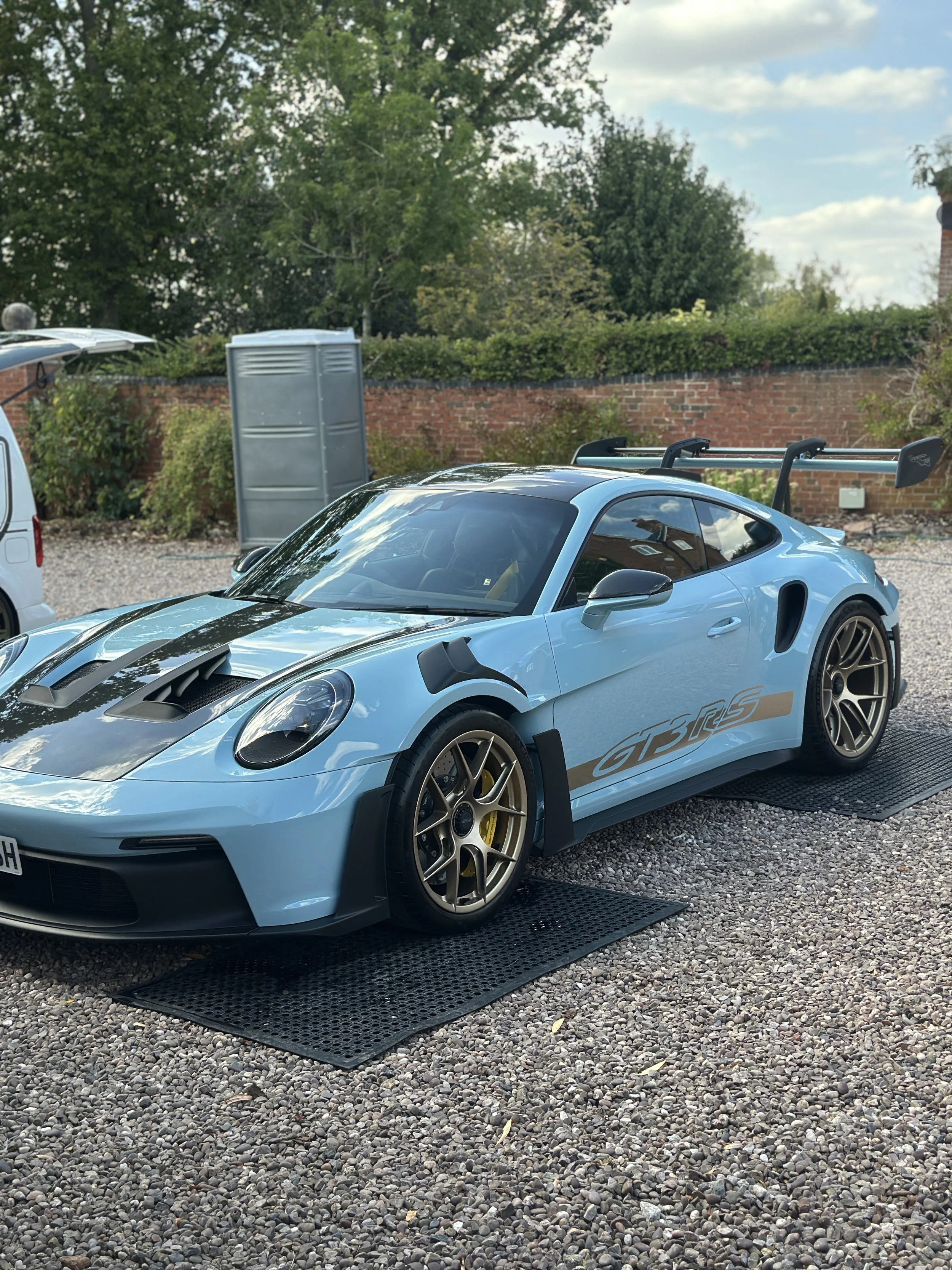 Light blue Porsche GT3 RS sports car with black stripes and a large rear wing, parked on a gravel lot with a black rubber mat underneath, in front of a brick wall and green trees.