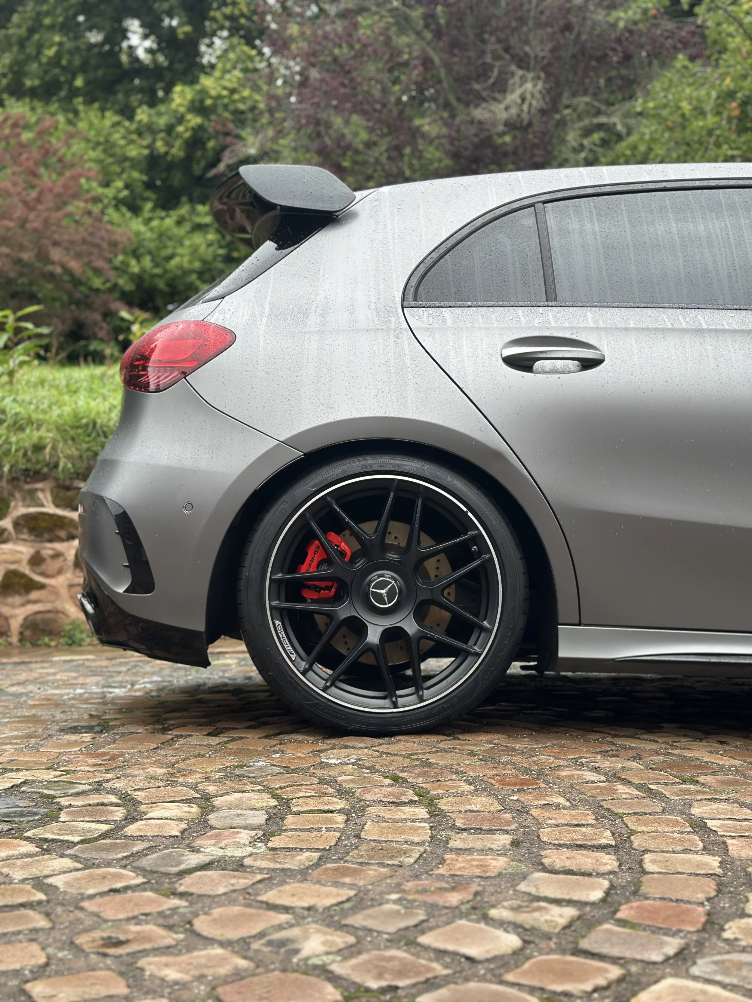 Close-up of the rear side of a gray Mercedes-Benz car parked on a cobblestone path with wet surface, showing the back and part of the side of the vehicle, with trees in the background.