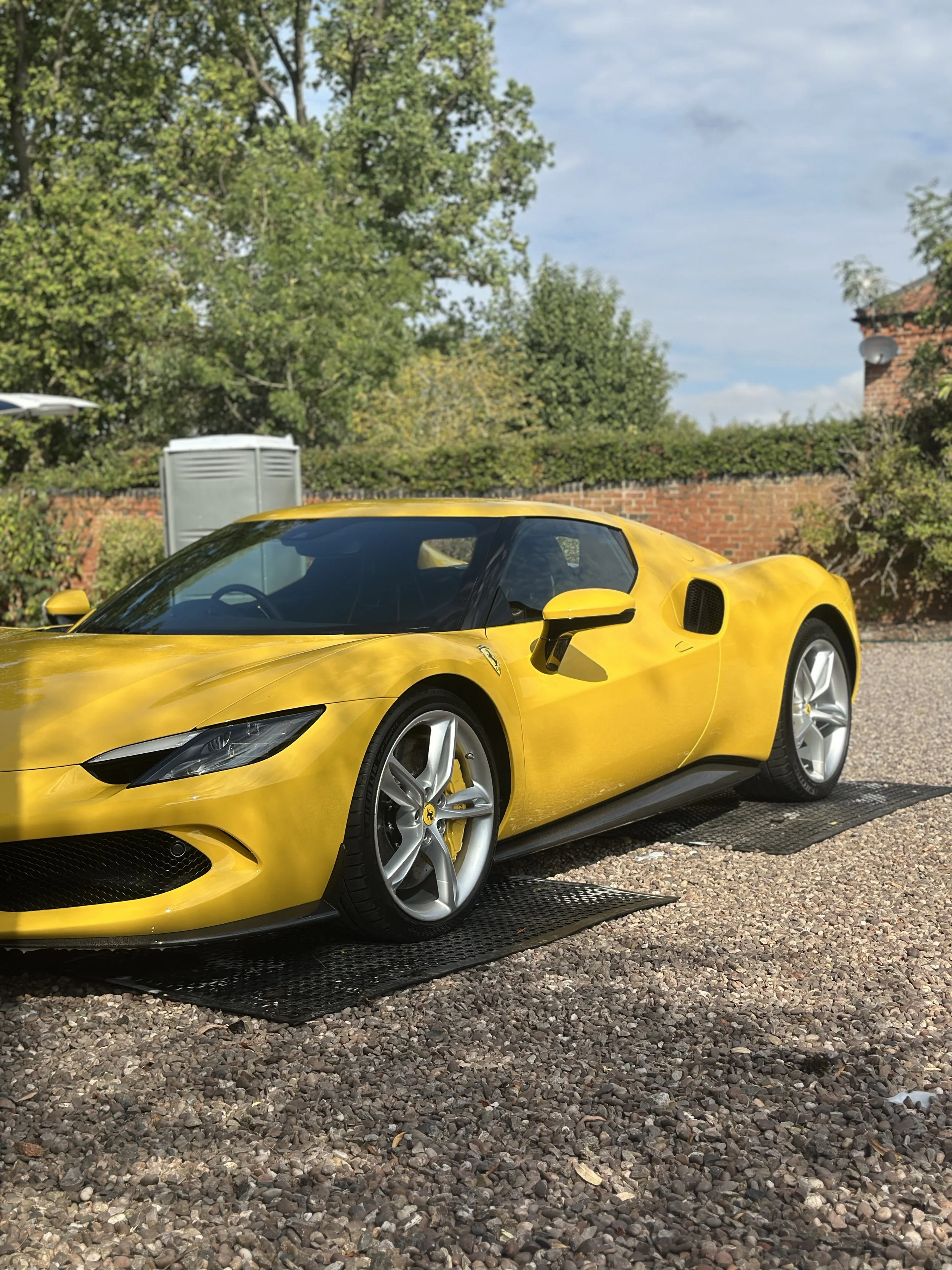 A bright yellow Ferrari 296 GTB, parked on a gravel surface under a partly cloudy sky, with green trees, bushes, and a brick wall in the background.