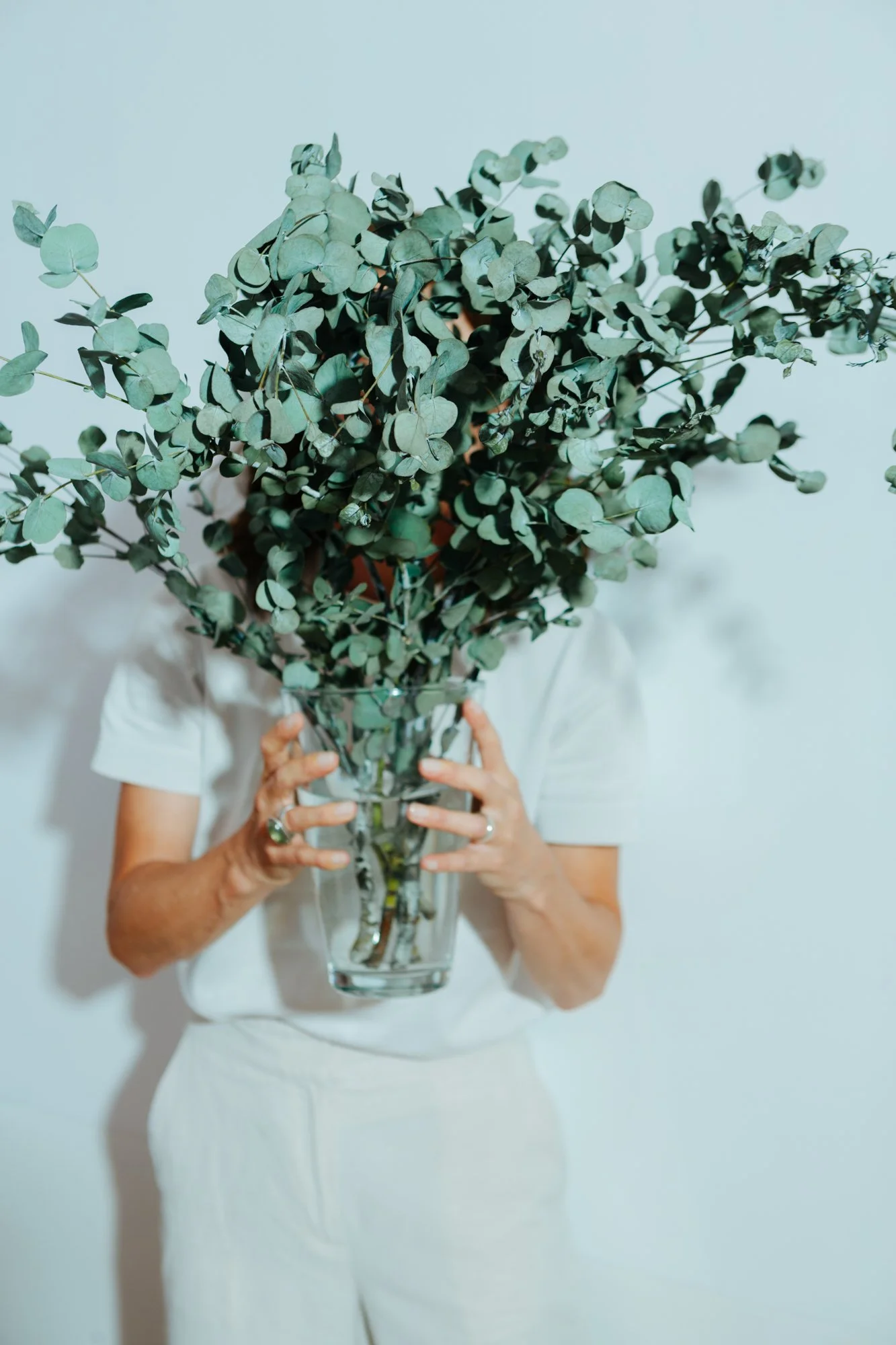 Eine Person hält eine Vase mit grünem Eukalyptus, die das Gesicht bedeckt.