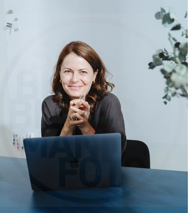 Eine Frau sitzt an einem Tisch vor einem Laptop, lächelt in die Kamera und hat ihre Hände vor ihrem Mund verschränkt, im Hintergrund ein Bücherregal und eine Pflanze.