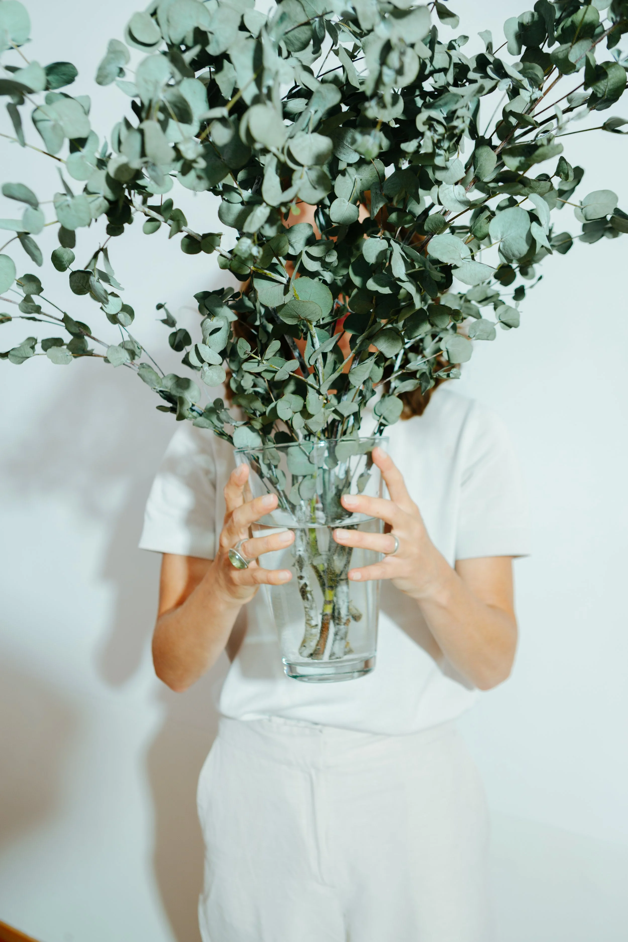 Eine Person in weißem Kleid hält eine Vase mit einem großen Zweig voller grüner Blätter vor ihr Gesicht.