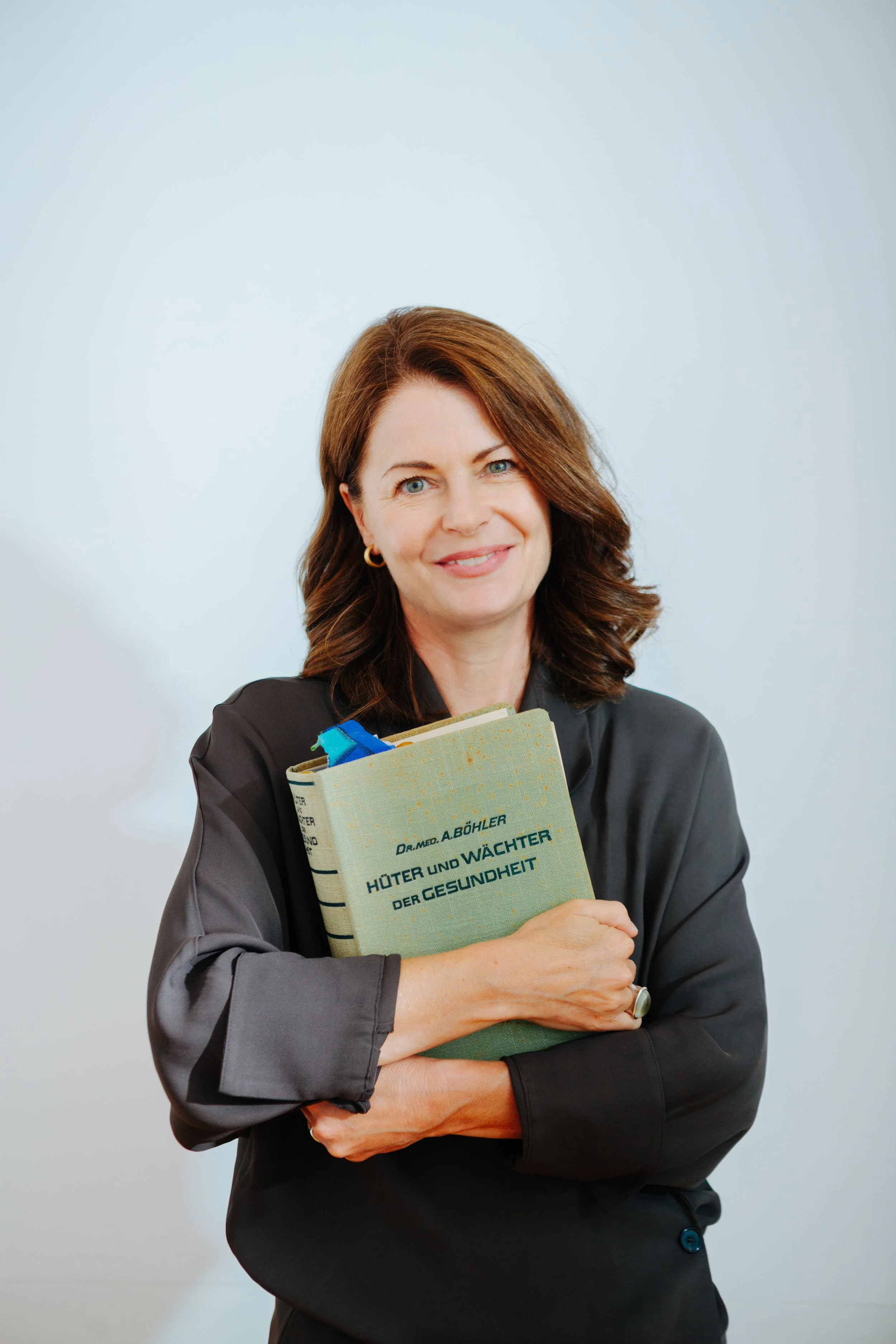 Eine Frau mit braunen, lockigen Haaren hält ein Buch mit einem grünen Cover vor ihrem Körper, trägt einen schwarzen Blazer und lächelt freundlich.