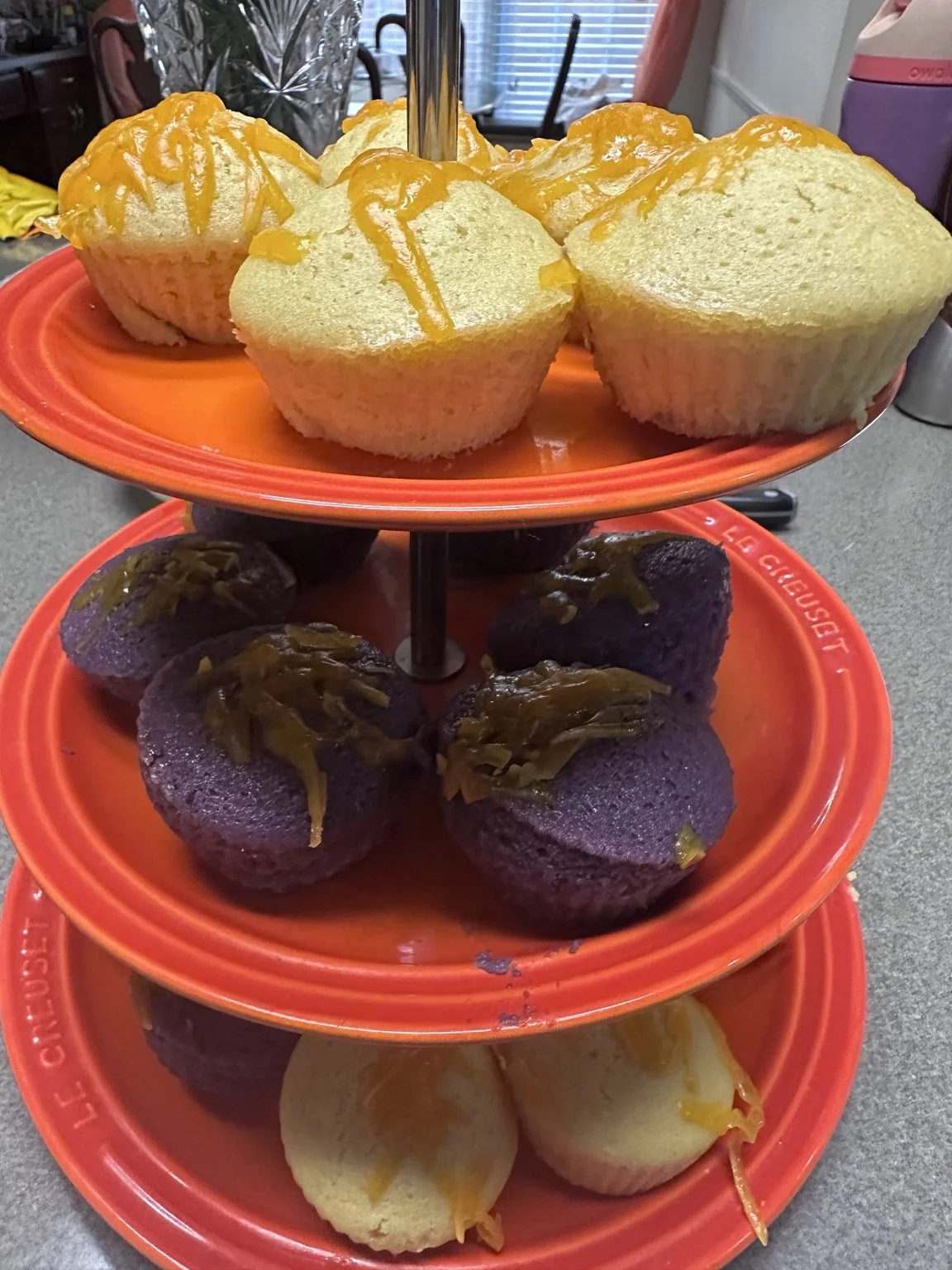 Yellow and ube Filipino puto topped with melted cheese on a tiered serving stand