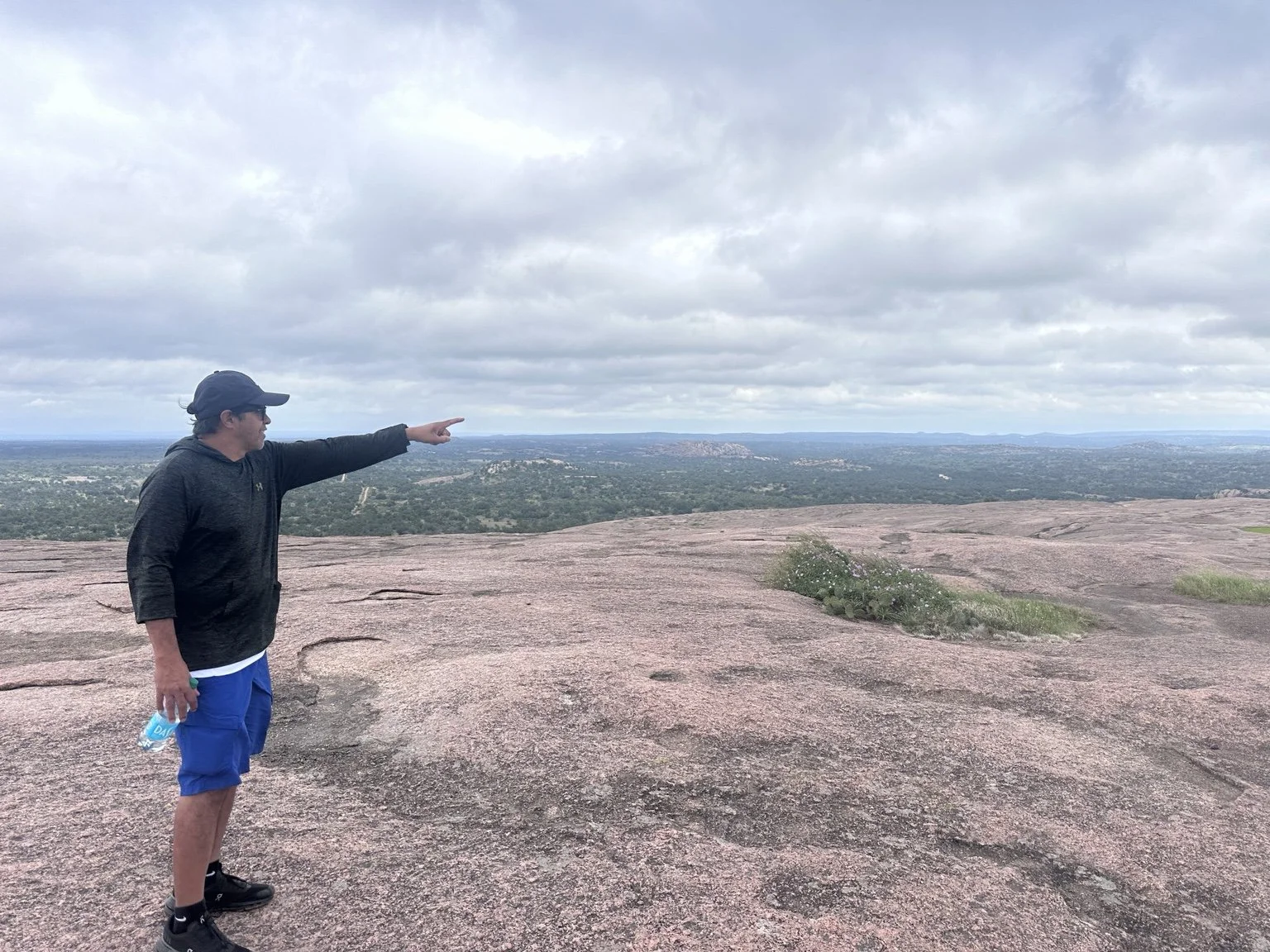 A hiker pointing across the broad pink granite slope with green Hill Country in the distance
