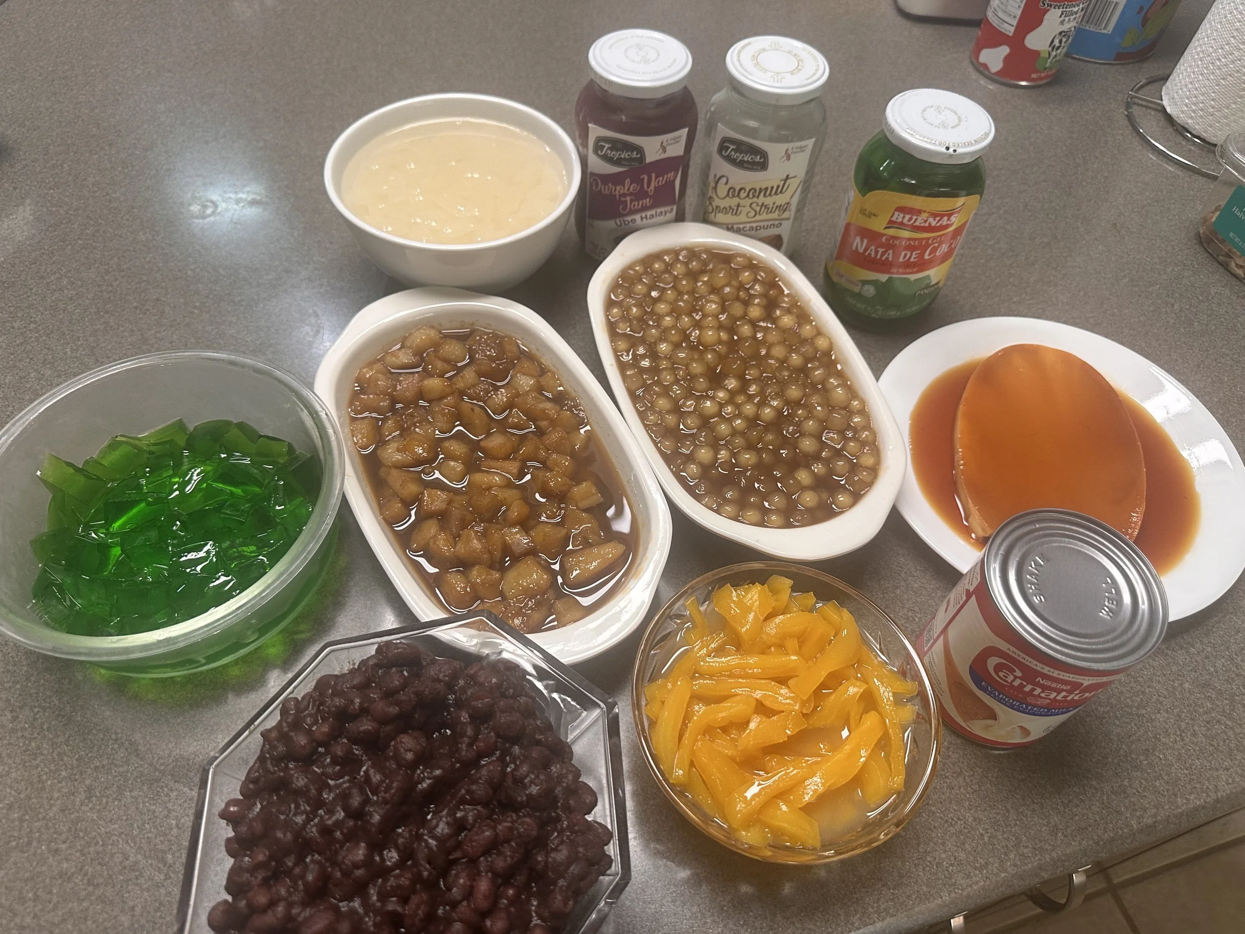 Halo-halo ingredients arranged on a kitchen counter, including green gulaman, sweetened fruits and beans, leche flan, and jars of additional toppings.