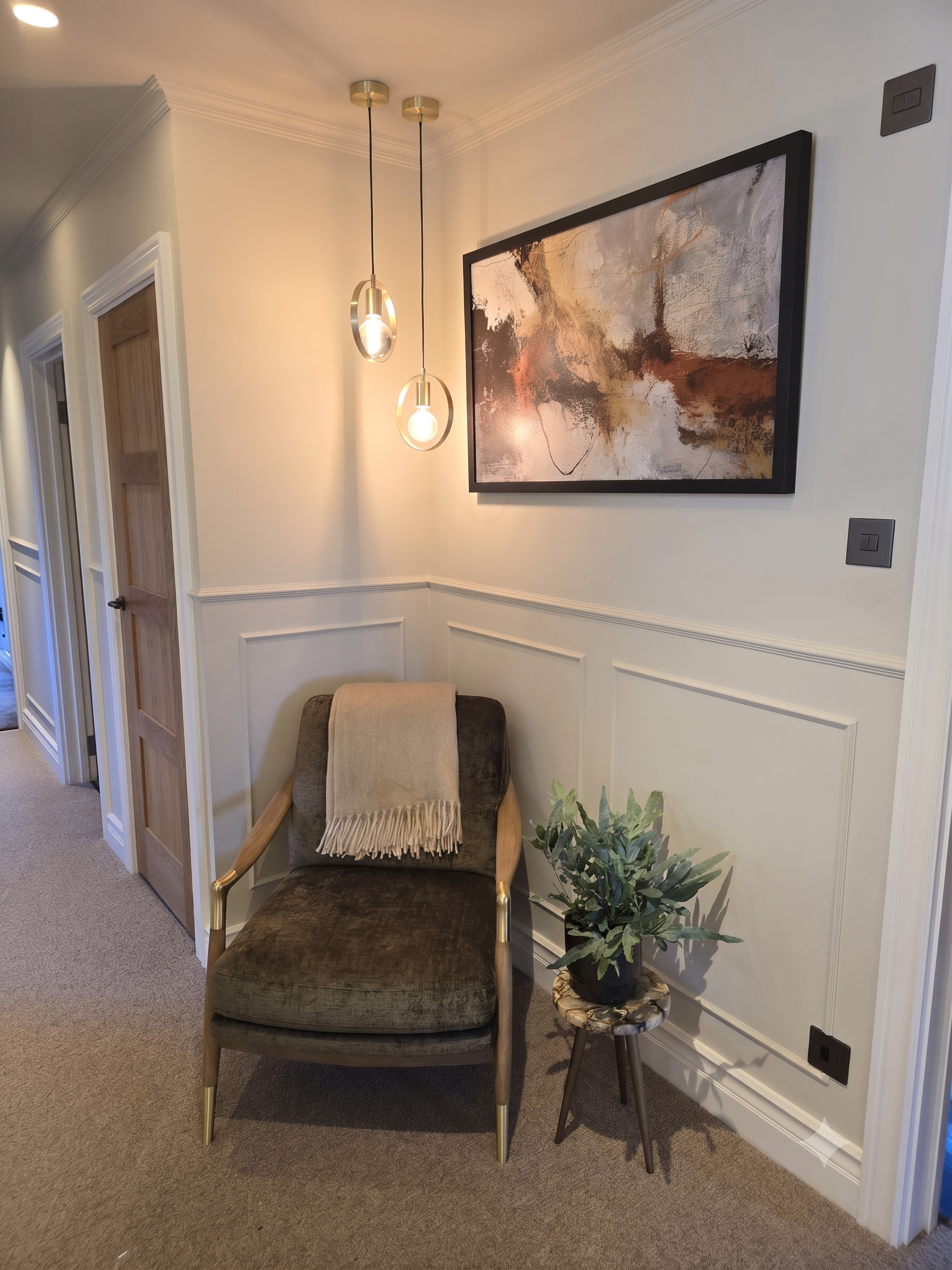 A cozy corner with a modern dark green upholstered armchair with wooden armrests. A beige throw blanket is draped over the back of the armchair. Next to the armchair, there is a small side table with a marble top and black metal legs, holding a potted green plant. The wall behind features white paneling and a large abstract painting with earthy tones. Two pendant lights with gold accents hang from the ceiling, and there are black power outlets on the wall.