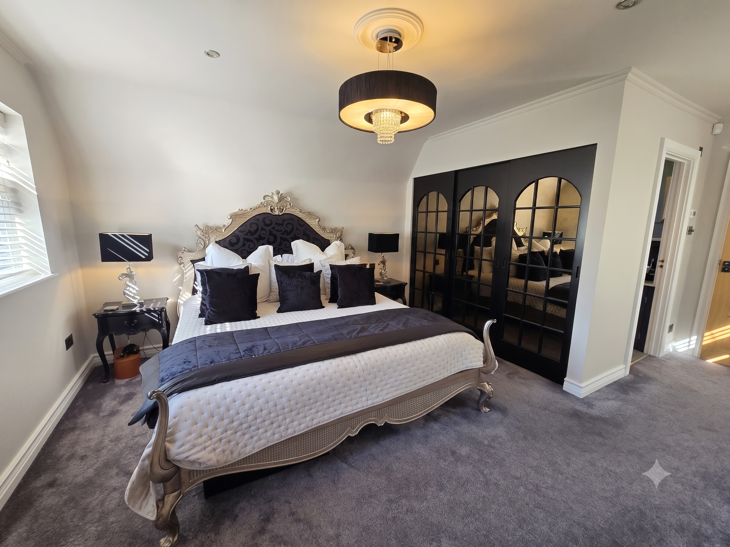 Elegant bedroom with a vintage silver bed frame, dark velvet bedding, multiple pillows, black lamps, and a modern black chandelier with crystal accents.