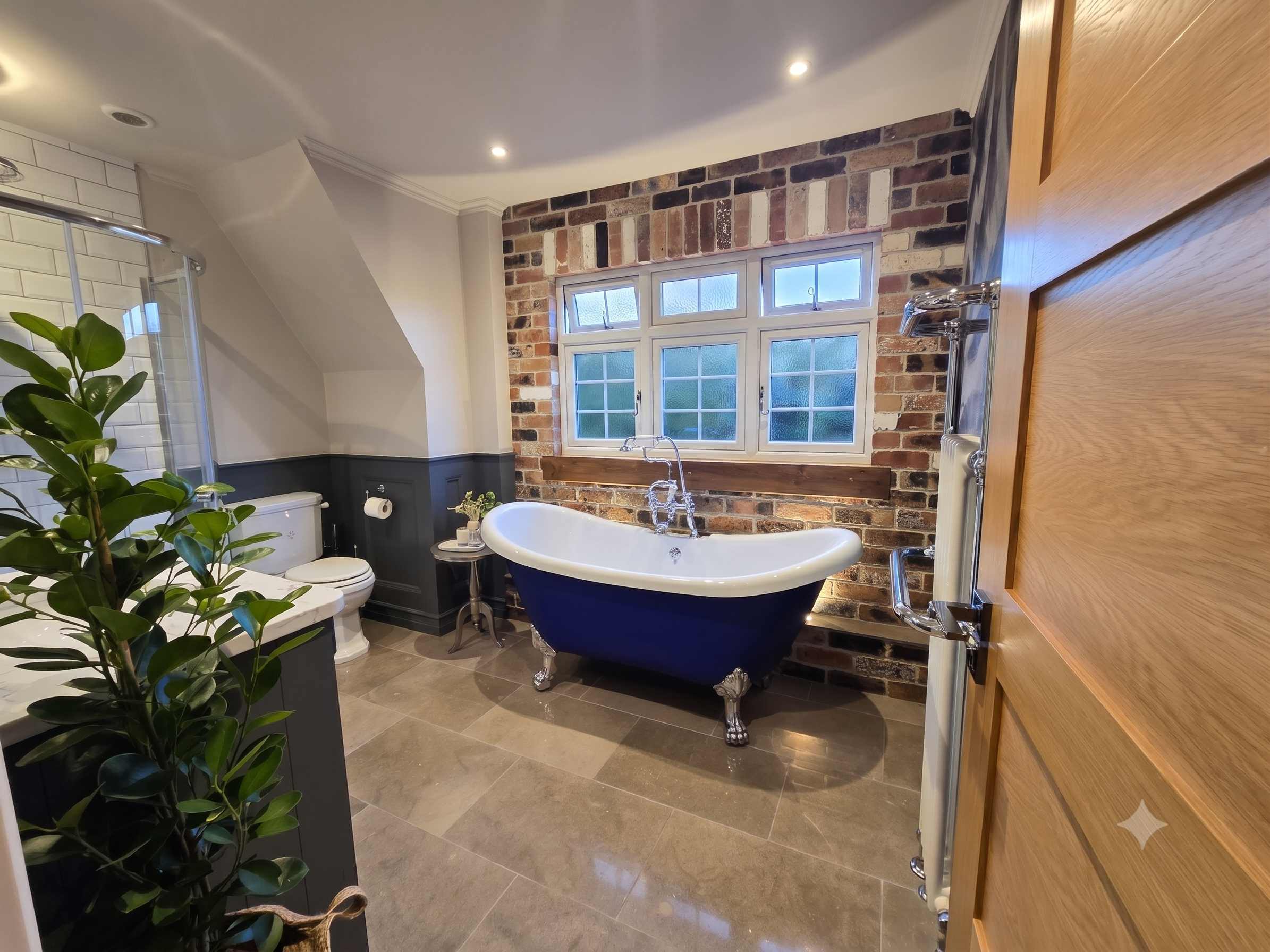 A bathroom featuring a clawfoot bathtub with a dark exterior and white interior, positioned in front of a brick wall with a window, and a wooden cabinet on the right side. There is a green potted plant in the foreground on the left, white subway tile