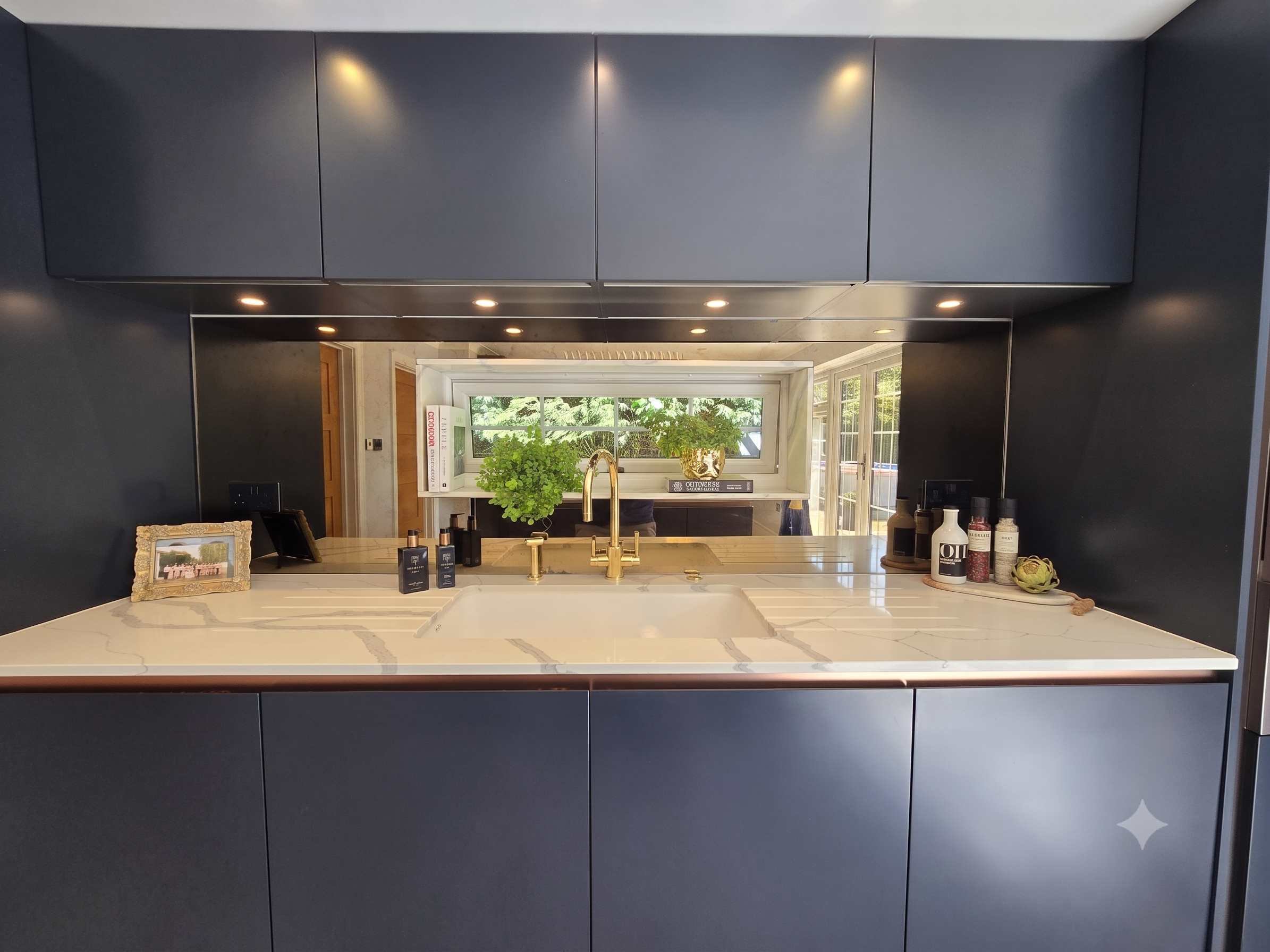 View of a modern kitchen with dark blue cabinets, a marble countertop, a gold faucet, and a window with a view of trees outside. The kitchen has potted plants, books, framed photos, and various bottles and containers on the counter.