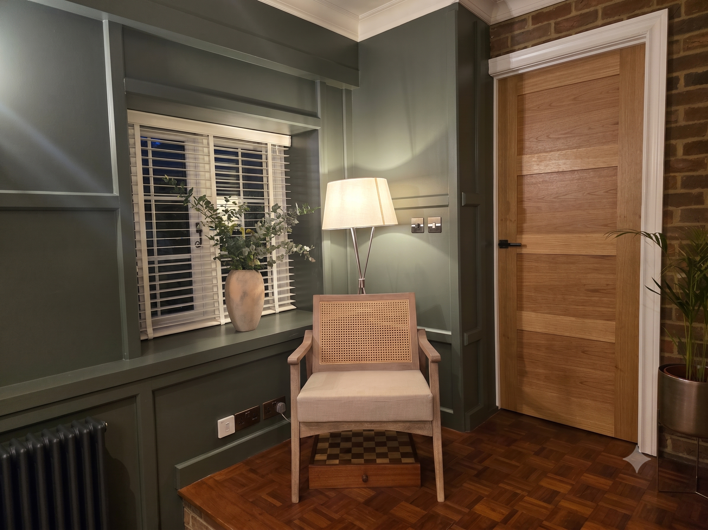 Interior room with green walls, a window with white blinds, a potted plant, a table lamp, a wooden door, a beige upholstered chair, and a corner with exposed brick wall.