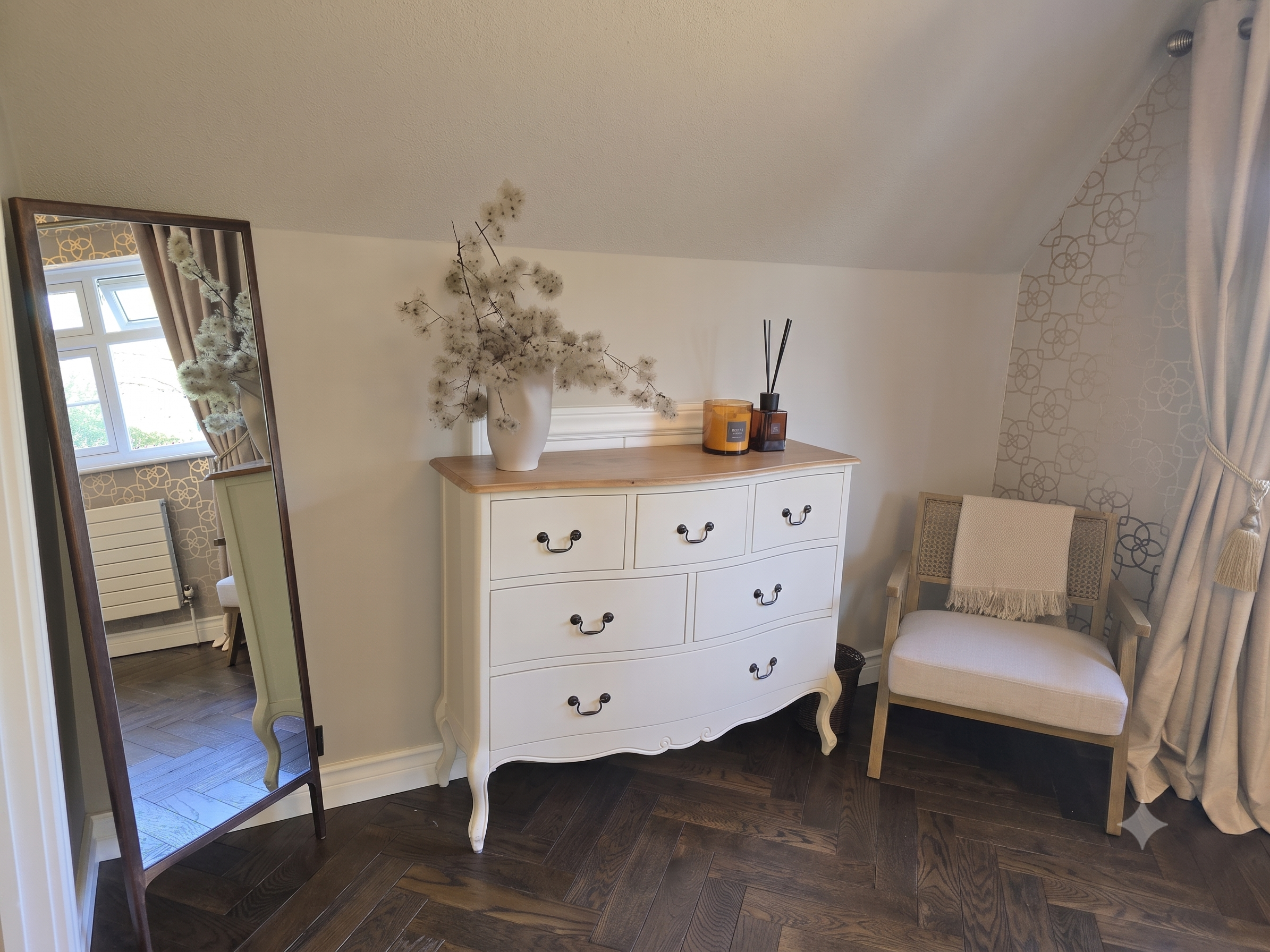 Interior of a room with a white dresser, a chair, and decorative items including a large vase with dried flowers, candles, a mirror, and curtains with a gold pattern. A window with curtains is reflected in a mirror.