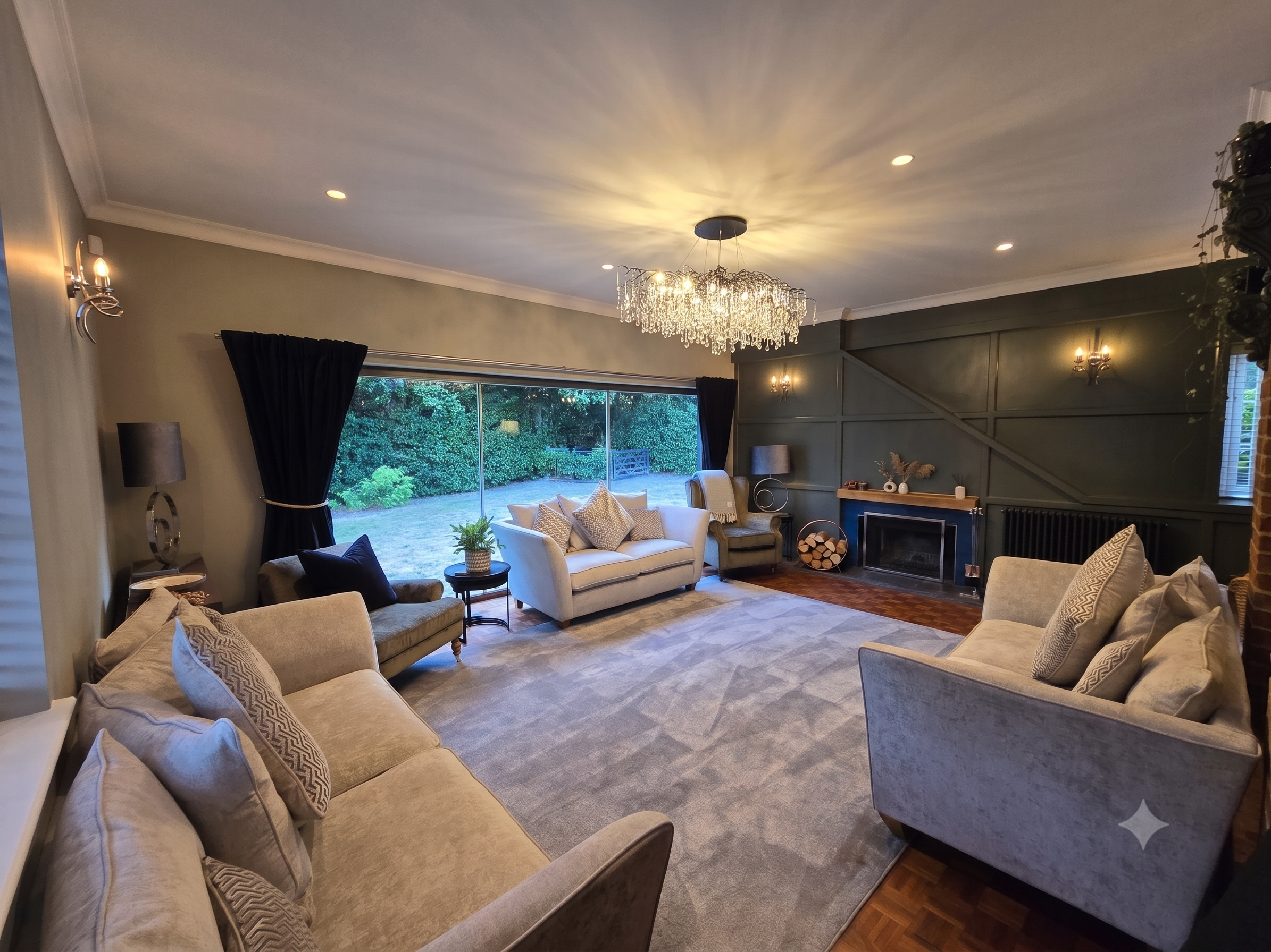 Living room with beige sofas, a fireplace, and a crystal chandelier, illuminated by wall sconces and lamps, with large window and green backyard view.
