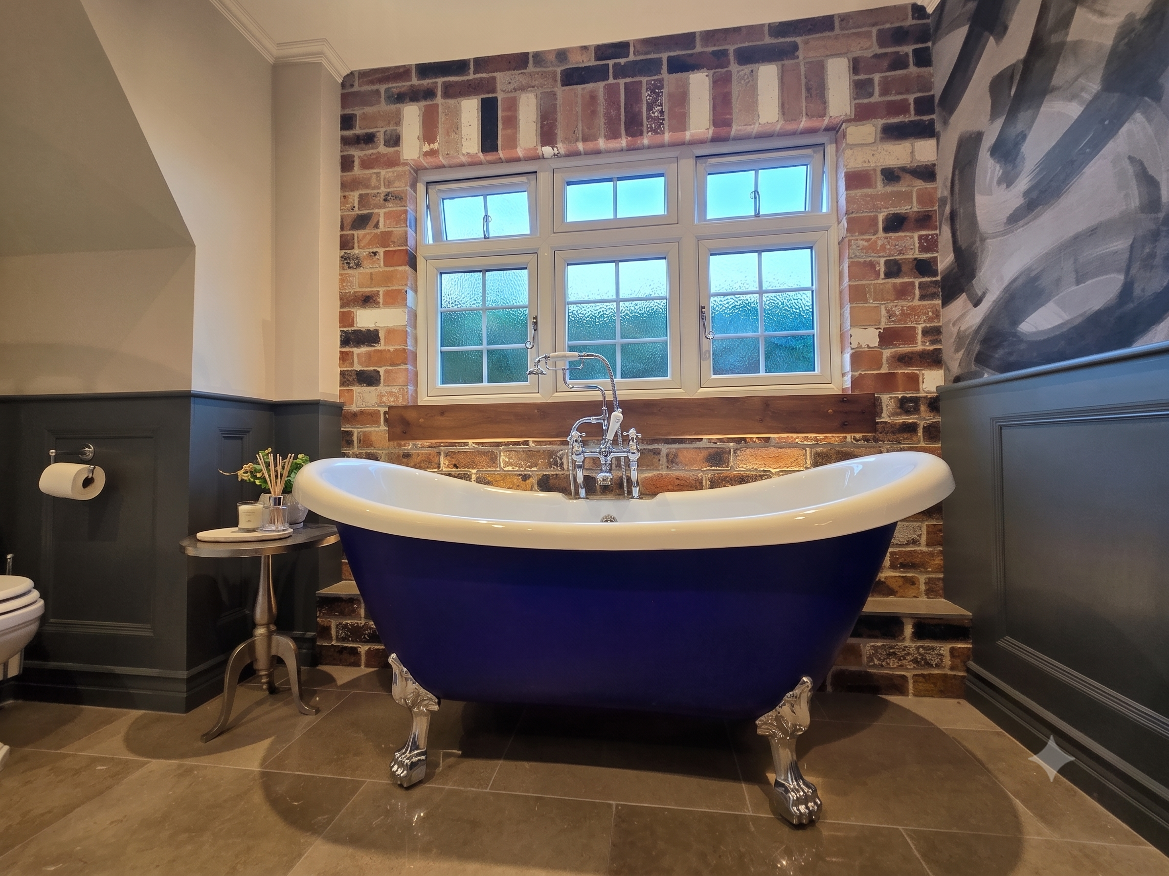 A bathroom with a freestanding clawfoot bathtub with a dark exterior and white interior, set against a brick wall and a large window. There is a small table with toiletries and a plant to the left of the bathtub, and the floor is tiled.