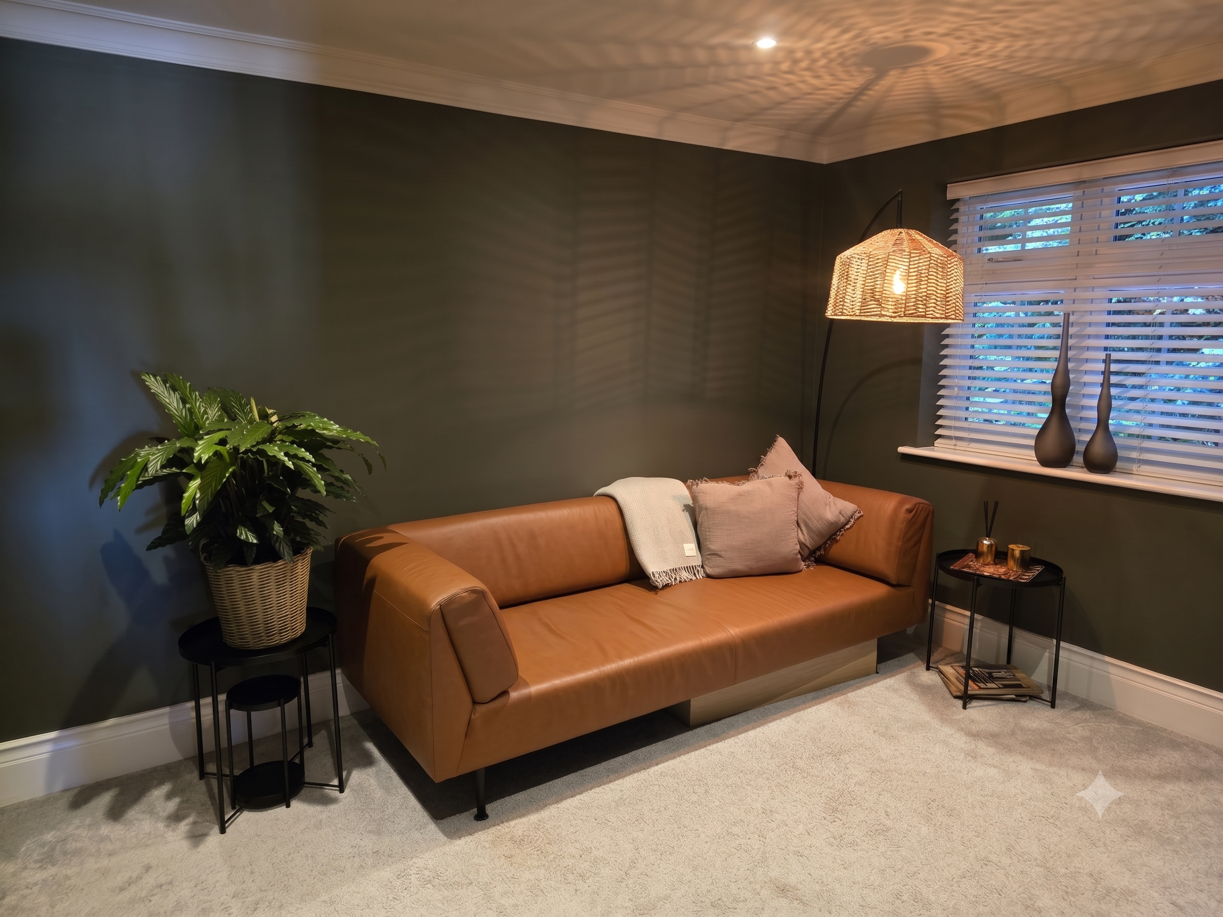 Living room with olive green walls, a leather sofa with pillows, a side table with decor, a tall potted plant, a floor lamp, window with white blinds, and a ceiling light pattern.
