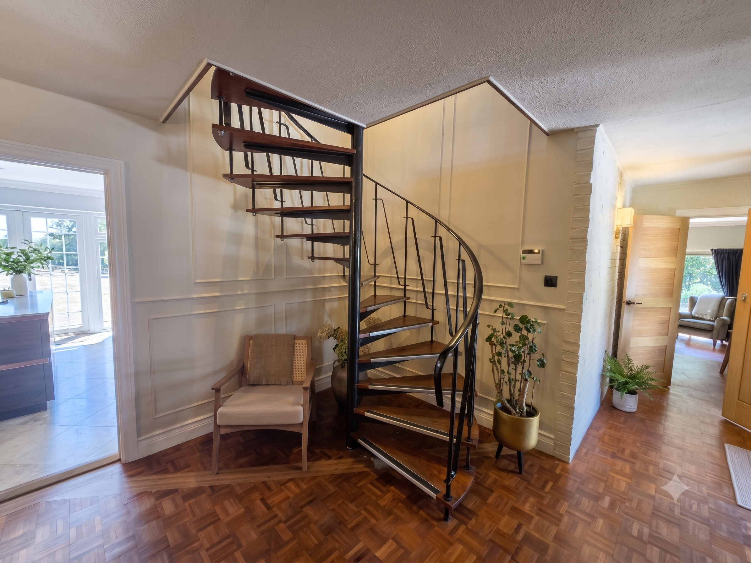 Interior view of a home showing a black spiral staircase with wooden steps, a beige armchair, potted plants, and a large window with a view outside.