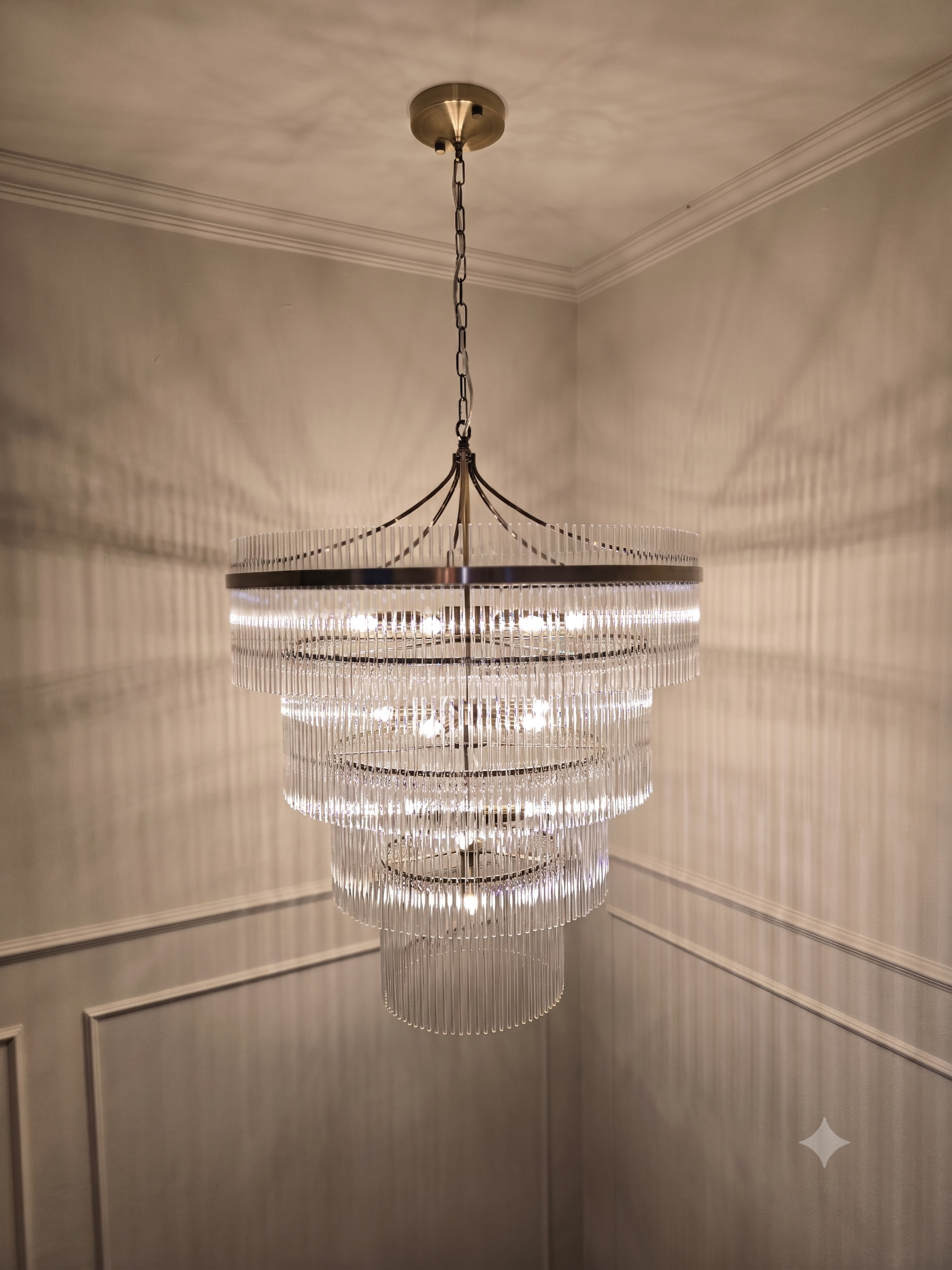 A modern chandelier with cascading circular tiers of vertical glass or acrylic rods hanging from a metal frame, suspended by a chain from a ceiling.
