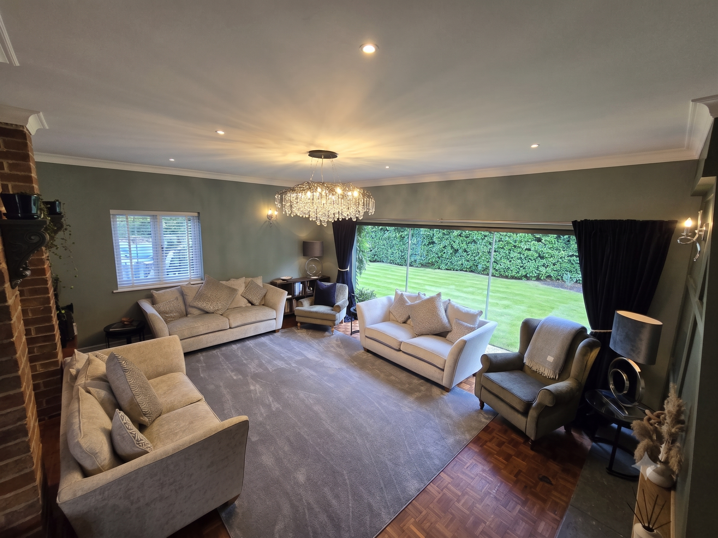 Living room with beige sofas, gray armchairs, a large window with dark curtains, a chandelier, and a view of a grass lawn and trees outside.