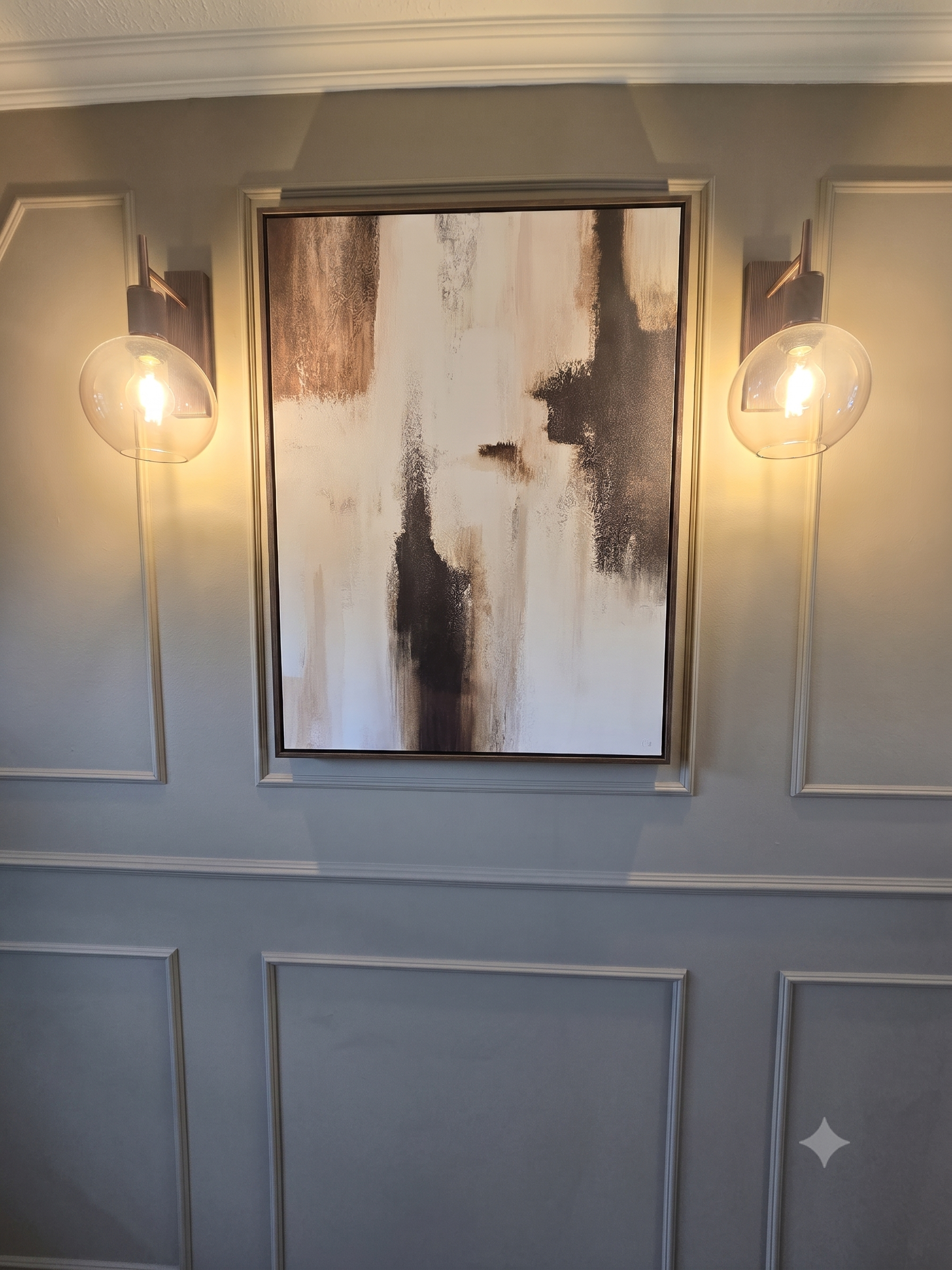 A framed abstract painting hangs on a beige wall with decorative white molding. Two modern wall-mounted light fixtures with glass globes are on either side of the painting, illuminating the artwork.