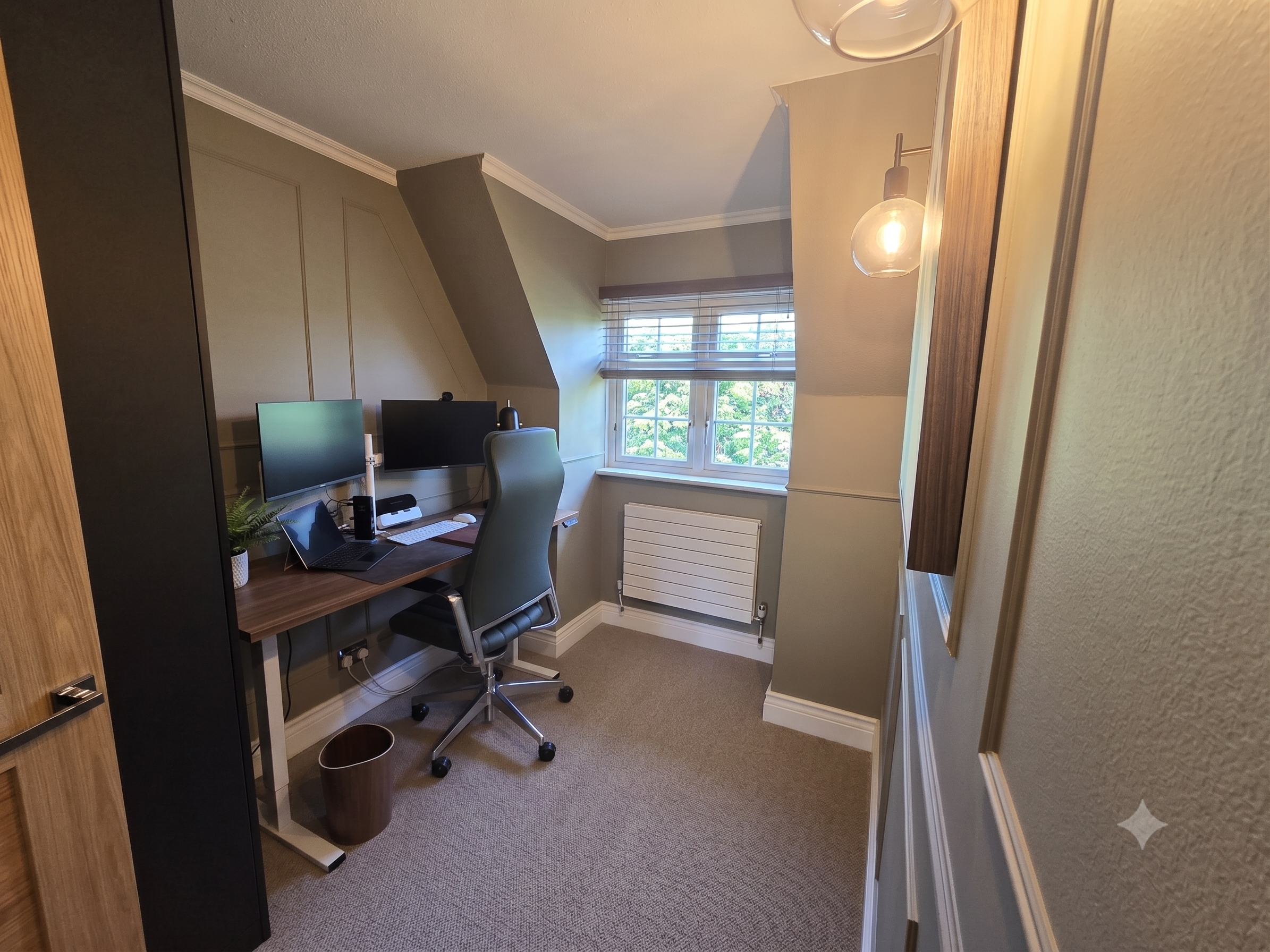 A small home office with a desk, computer monitors, a chair, a window with blinds, and a wall-mounted light fixture.