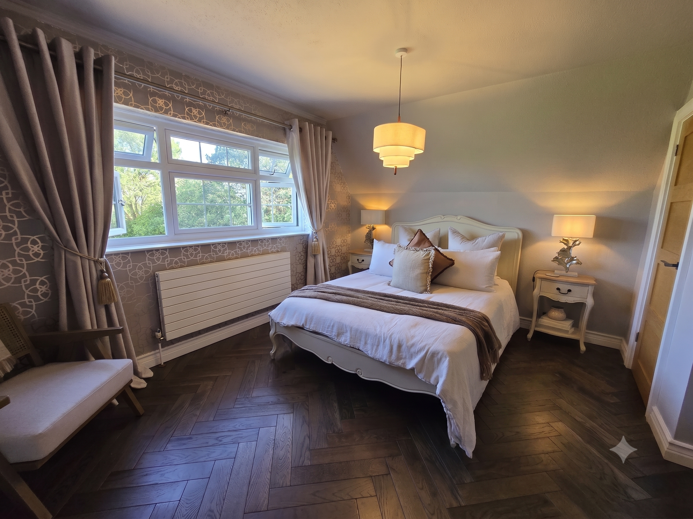 A cozy bedroom with a large window, beige curtains, a white bed with pillows, two bedside lamps, a table lamp, and hardwood flooring.