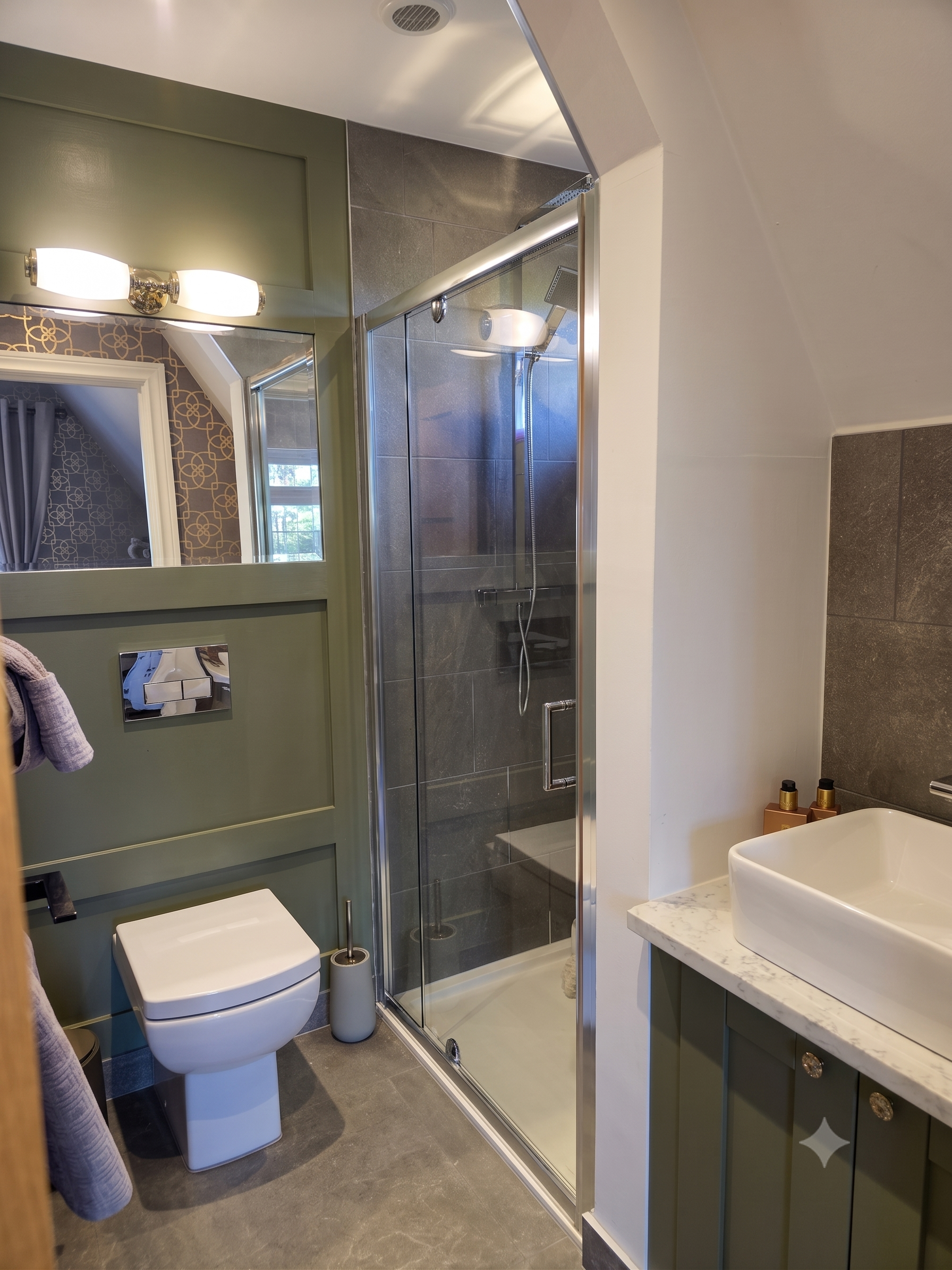 A modern bathroom with a glass shower enclosure, a wall-mounted toilet, a white rectangular vessel sink on a marble countertop, and green cabinetry with decorative knobs. There is a mirror above the sink and wall-mounted light fixtures.