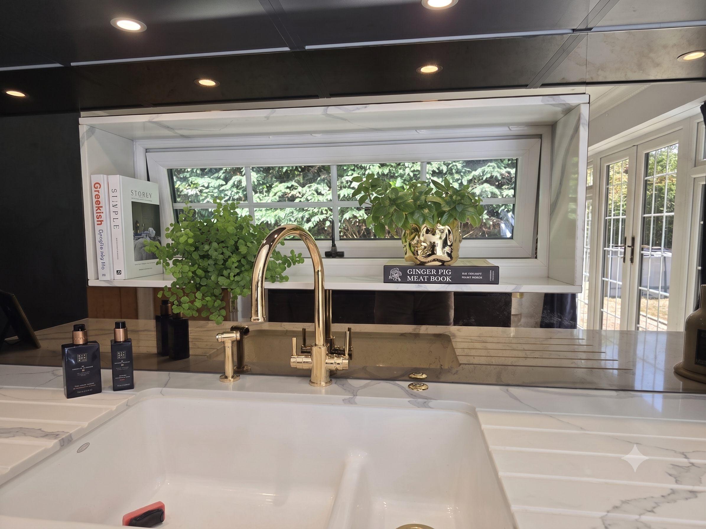 Kitchen countertop with two black bottles, green plants, a book titled 'Ginger Pig Meat Book', a window with greenery outside, and a gold-colored faucet.