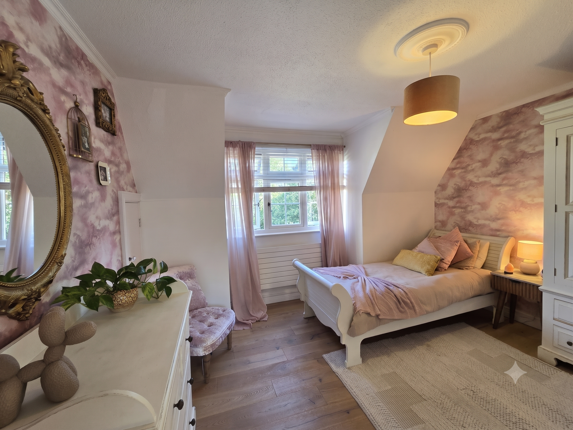 A cozy bedroom with a white bed, pink and beige pillows, soft pink curtains, a large window, a vanity table with decorative objects, a mirror with an ornate gold frame, and a pink themed accent wall with clouds.