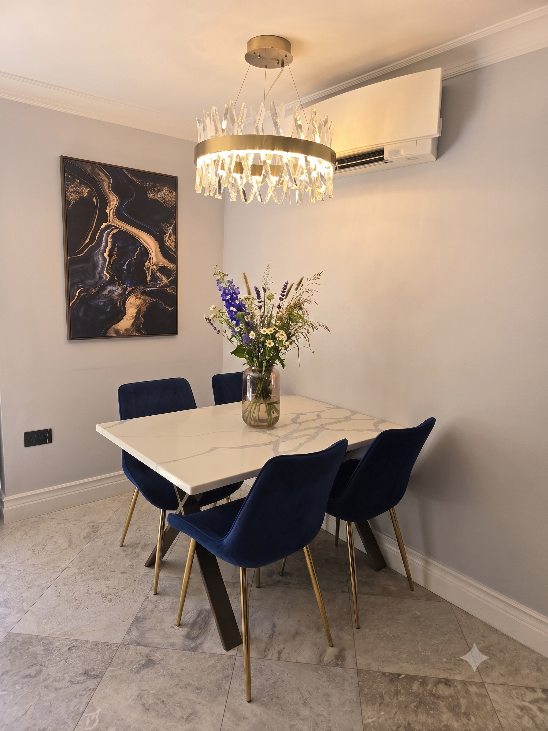 A modern dining room with a white marble table, four navy blue chairs with gold legs, a vase with purple and white flowers, a abstract black and gold wall art, a crystal chandelier, and wall-mounted air conditioning.