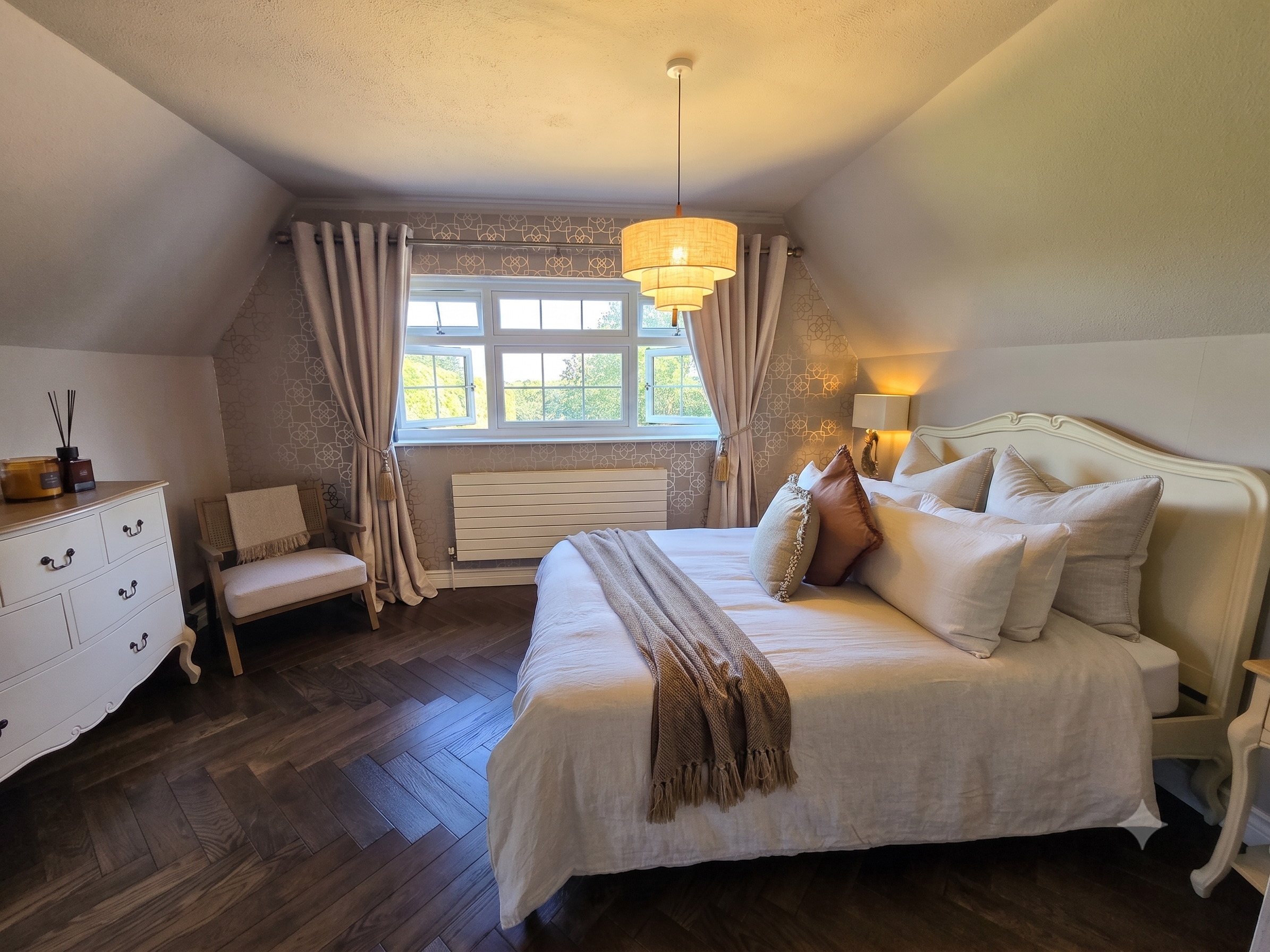 A cozy bedroom with a large bed covered in light-colored bedding and pillows. There is a bedside table with a lamp and a chair next to a window with beige curtains. The room has sloped ceilings with a hanging pendant light.