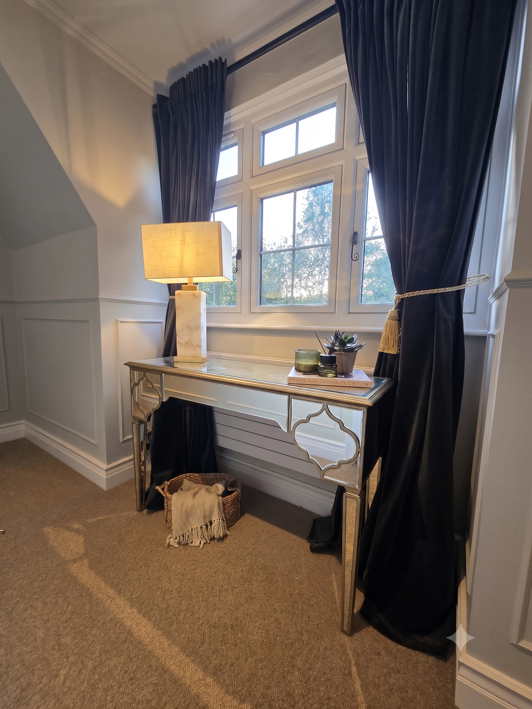 A decorative console table with a marble top, a lamp, a potted succulent, and a couple of books, situated under a window with dark navy curtains tied back. A woven basket with a blanket is on the floor next to the table.