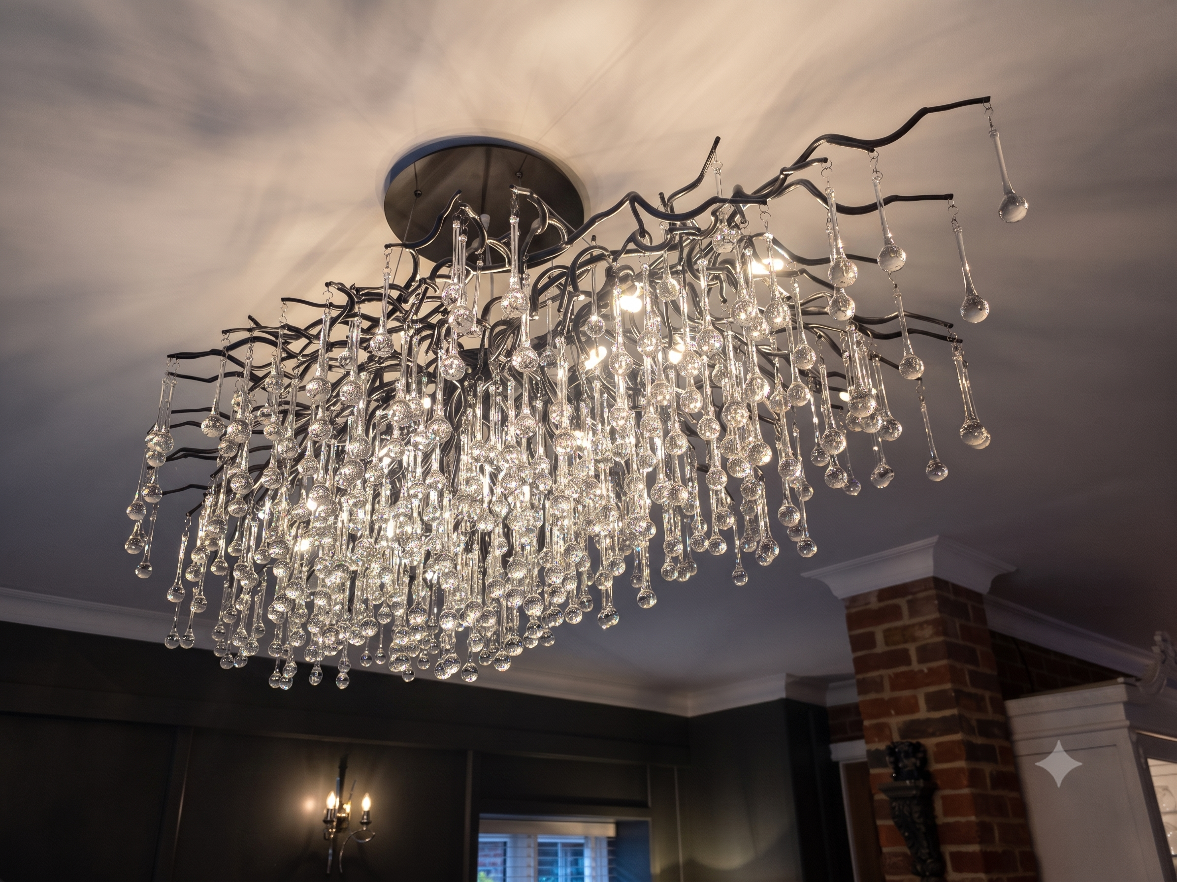 A modern chandelier with glass droplet pendants hanging from curved metal arms, mounted on a ceiling in a room with brick wall accents.