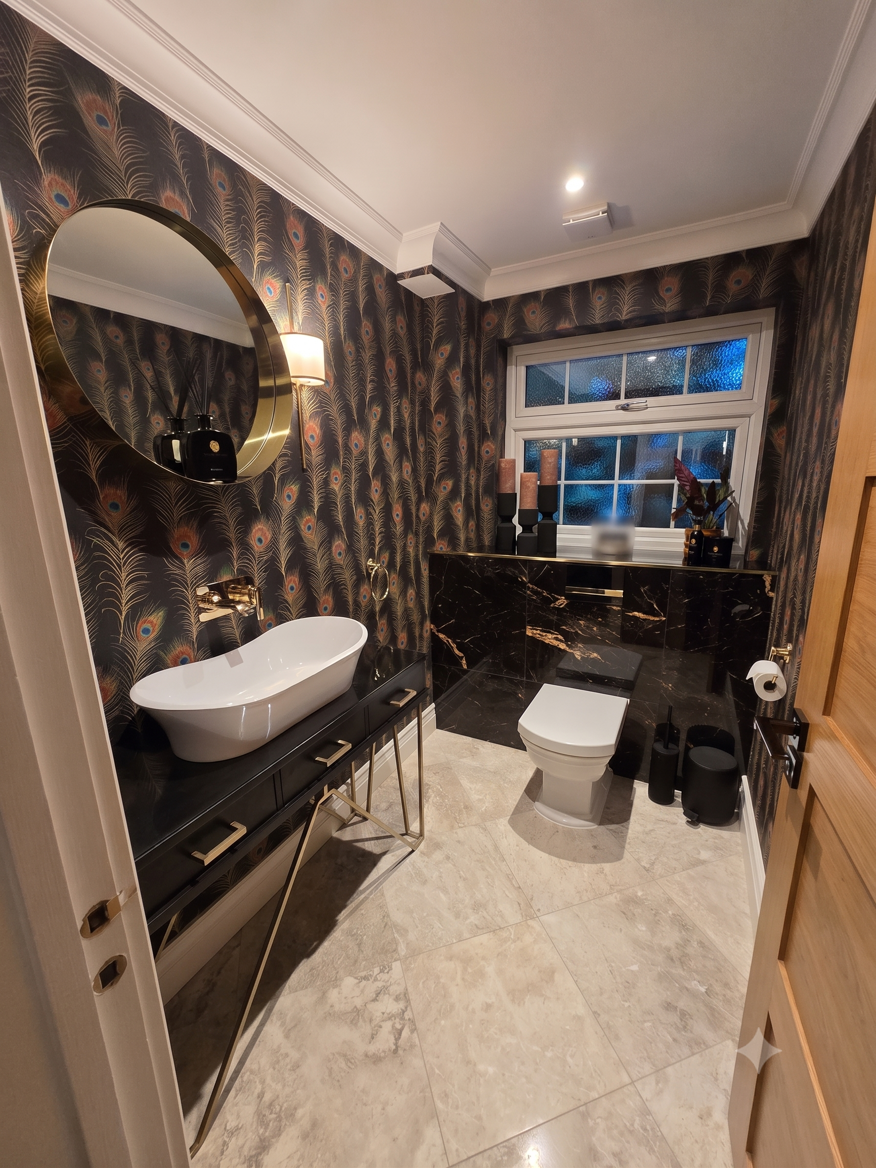 A bathroom with black marble vanity, a white bathtub, a toilet, decorative peacock feather wallpaper, and a window with textured glass.
