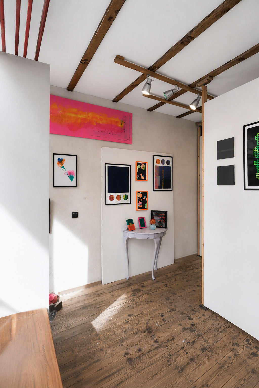 Interior view of an art gallery or studio with various colorful framed artworks on white walls, a small antique white table with decorative items, and a wooden ceiling with track lighting.