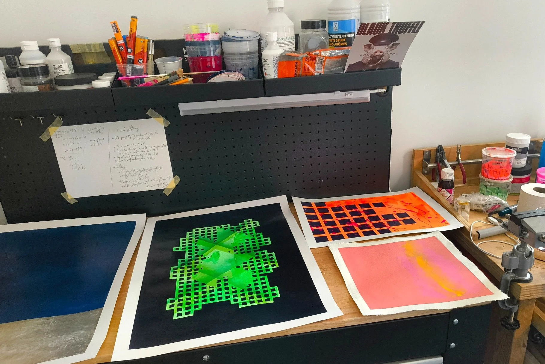 Workstation with art supplies, printed colorful designs, and art tools on a desk and pegboard, including paint, markers, and printed digital art images.