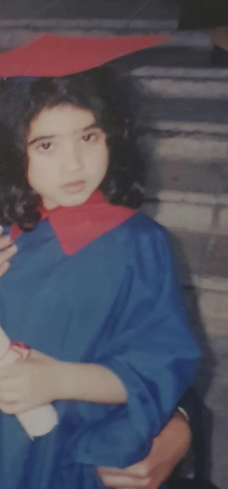 Young girl in a graduation gown with a red collar and blue robe, holding a diploma.