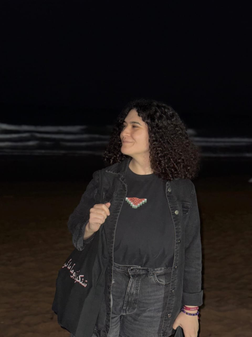 A young woman with shoulder-length curly hair standing on a dark beach at night, smiling and looking to the side, wearing a black jacket and a black shirt with a pixelated watermelon graphic.
