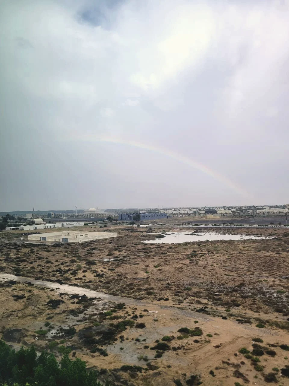 A landscape with arid land, some green bushes, a small lake, and several industrial buildings in the distance. A faint rainbow is visible in the cloudy sky.