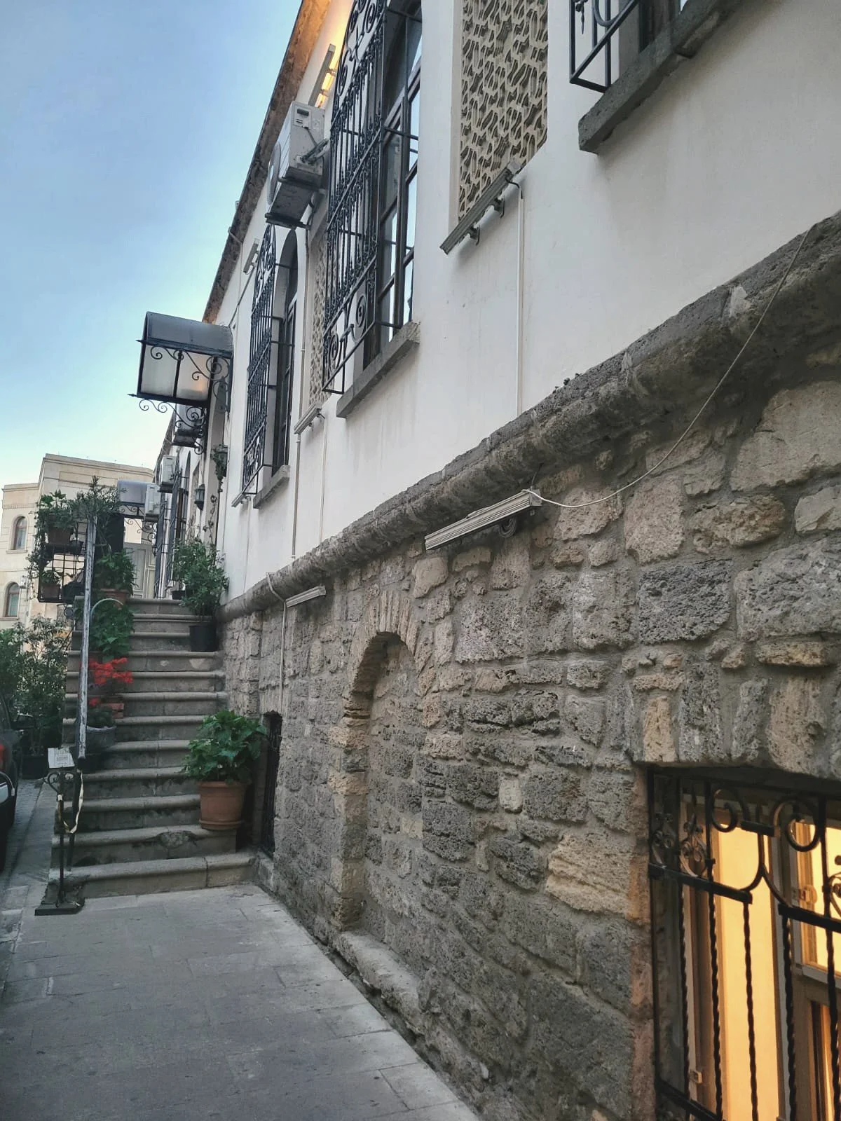 Stone building with stairs leading to the entrance, windows with wrought iron bars, and plants on the steps, in an urban setting.