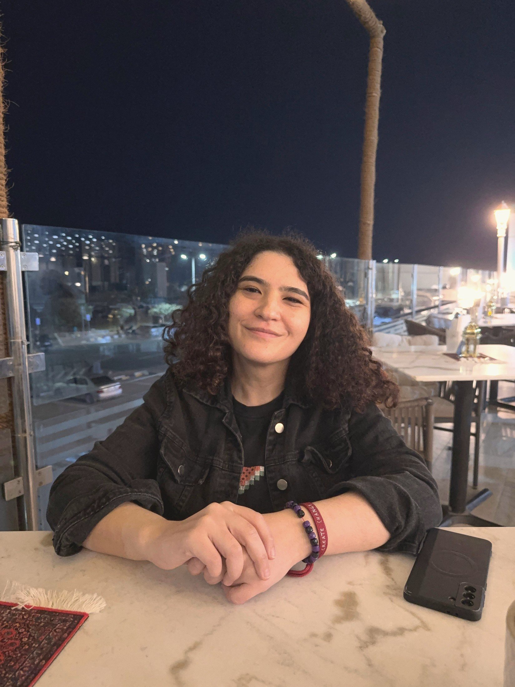 A young woman with curly hair smiling at the camera, sitting at a marble table with her hands clasped. She is wearing a black jacket and colorful bracelets on her wrist. The background shows a night scene with palm trees, a fence, parked cars, and street lights.