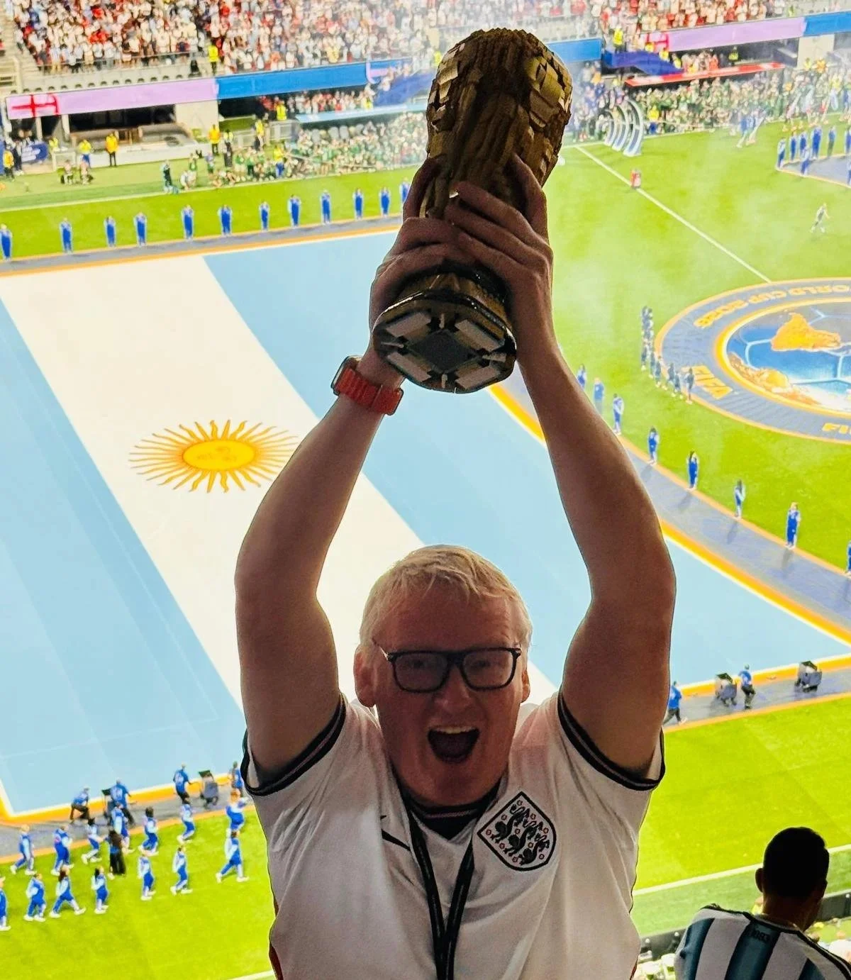 A man wearing glasses and a white England football jersey holding a replica World Cup trophy above his head at a football stadium, with the Argentine flag on the pitch and a crowd in the background.