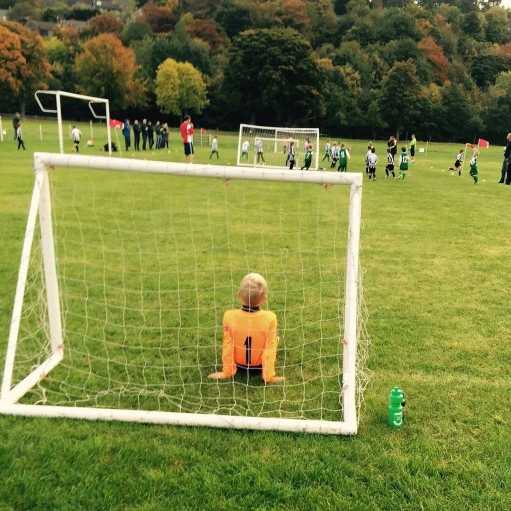 A young soccer goalie in an orange jersey sits in front of the net, watching a game on a grassy field. The game involves children and adults, with some children playing on the field and others standing nearby. The background shows trees with autumn foliage and a slightly overcast sky.