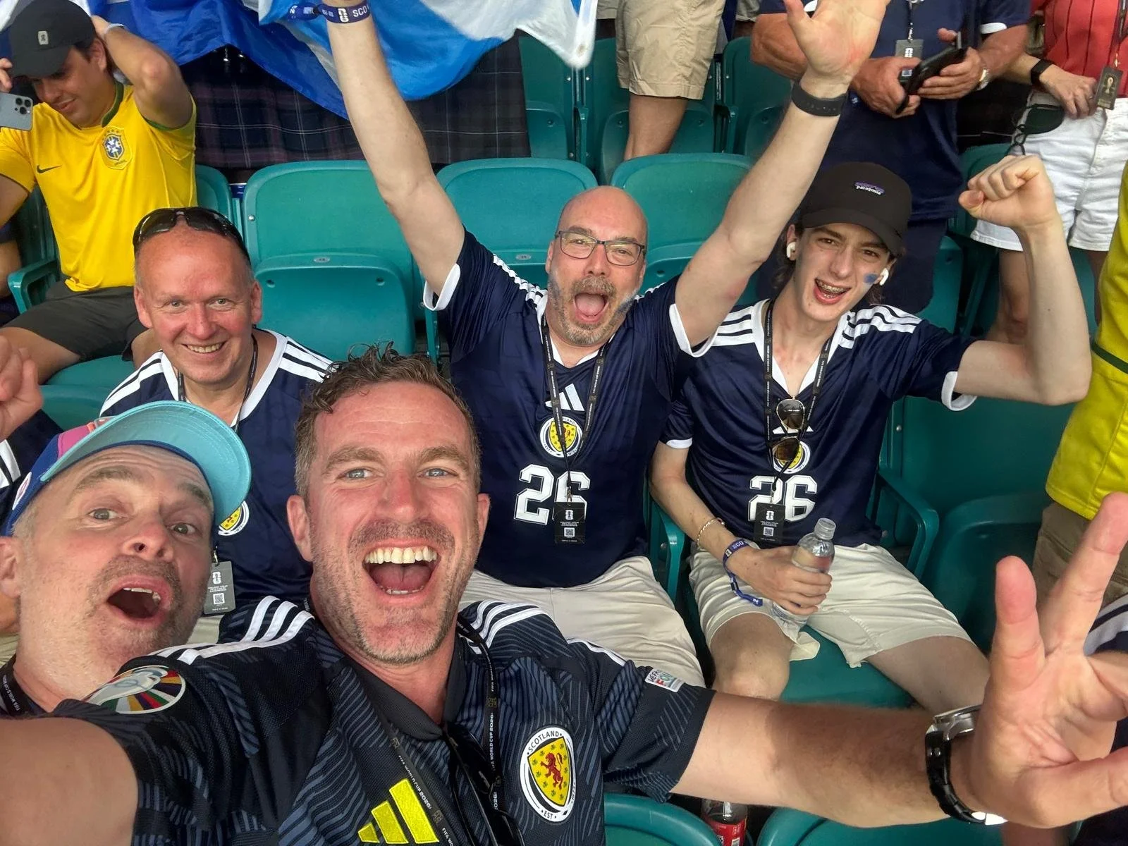 Group of people at a sports event, smiling and cheering, wearing Scotland national football team jerseys, some with face paint and accessories, seated in stadium seats.