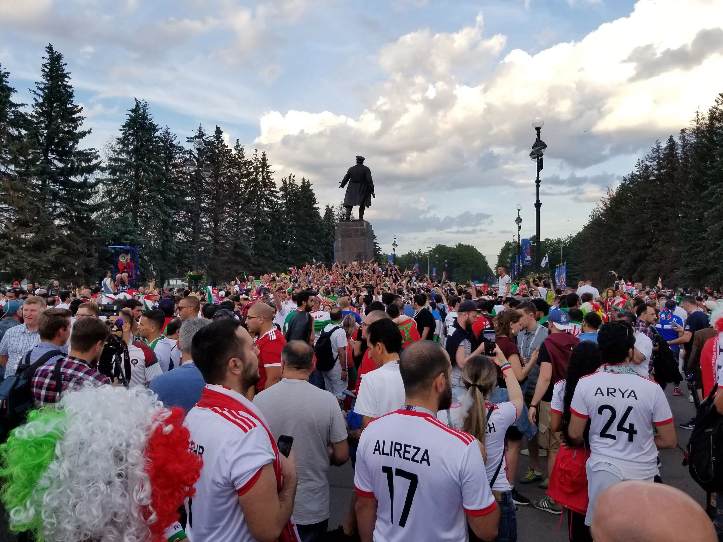 Crowd of football fans wearing jerseys, with one person dressed as a clown with a curly wig, gathered around a statue of a historical figure in a park with trees and cloudy sky in the background.