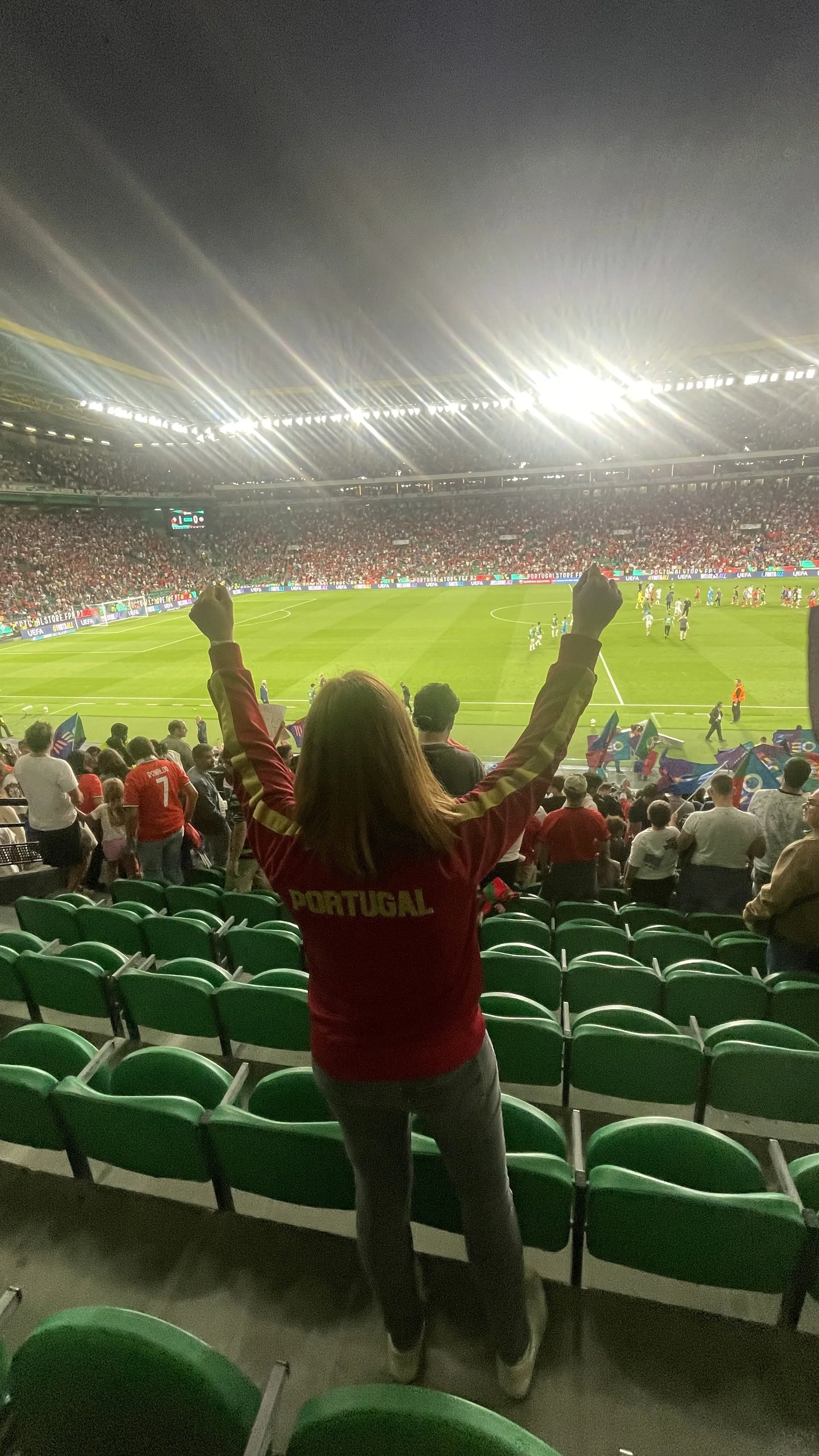 A woman wearing a Portugal soccer jersey standing with arms raised inside a large stadium filled with spectators, celebrating at the end of a Portugal versus Ireland football match.