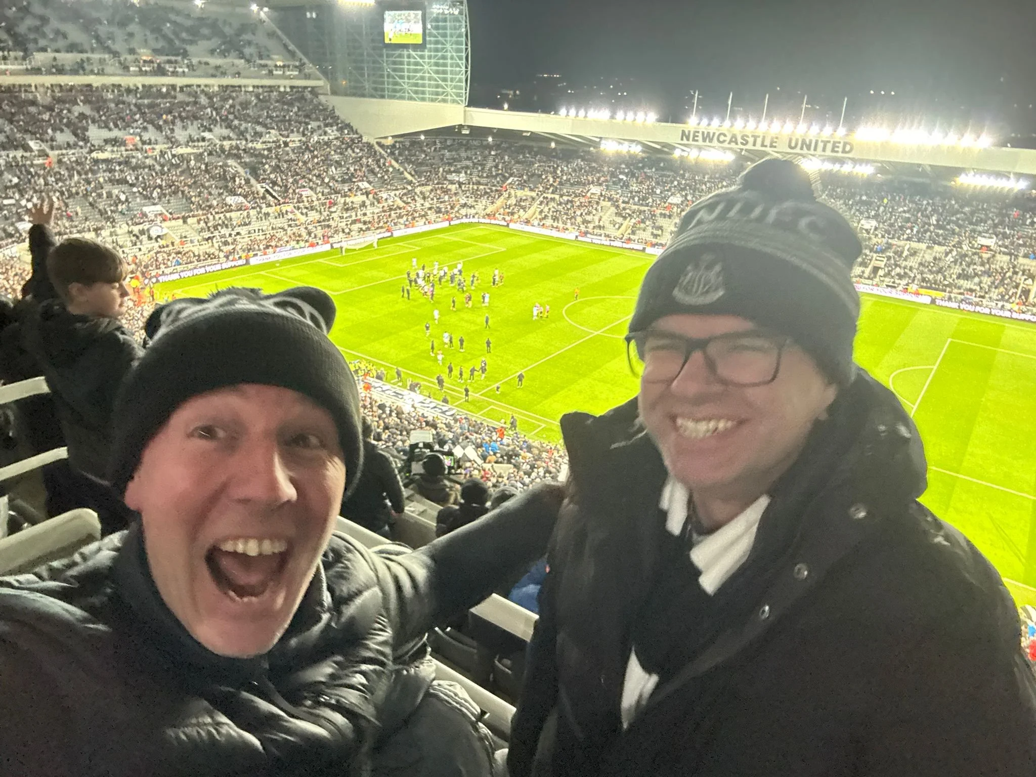 Two men in jackets smiling and taking a selfie at a night football match at Newcastle United stadium, with a view of the field and stands full of spectators in the background.