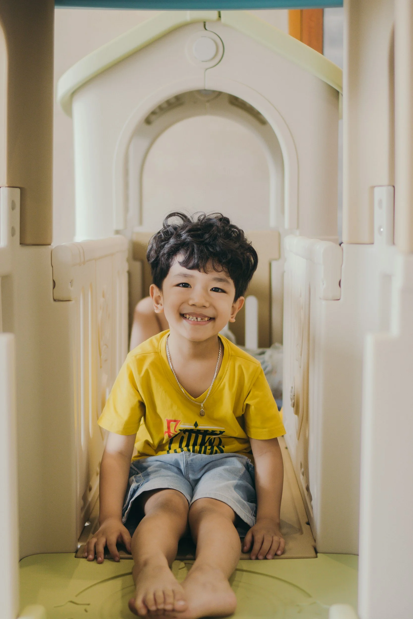 Un garçon souriant portant un t-shirt jaune est assis dans un toboggan pour enfants en plastique beige à l'intérieur, avec une structure de jeu en arrière-plan.
