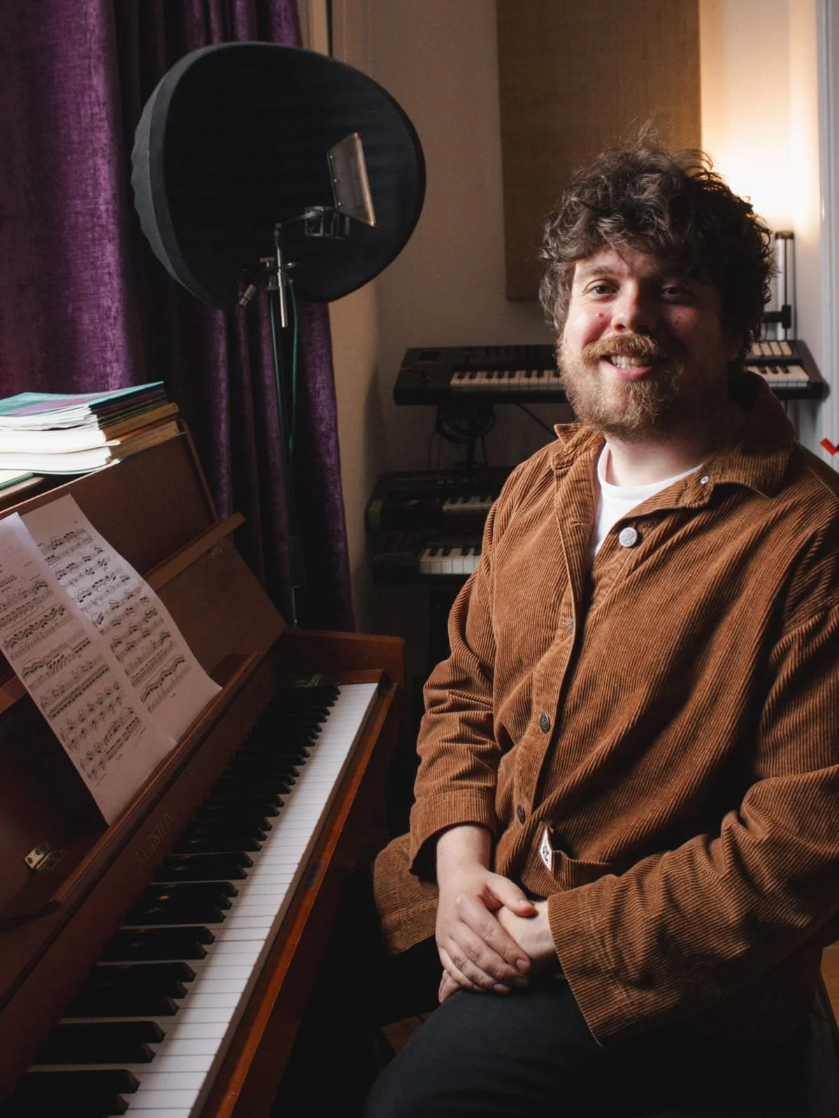 A man with curly hair and a beard smiling while sitting next to a wooden piano with sheet music on it, in a room with purple curtains and a keyboard in the background.